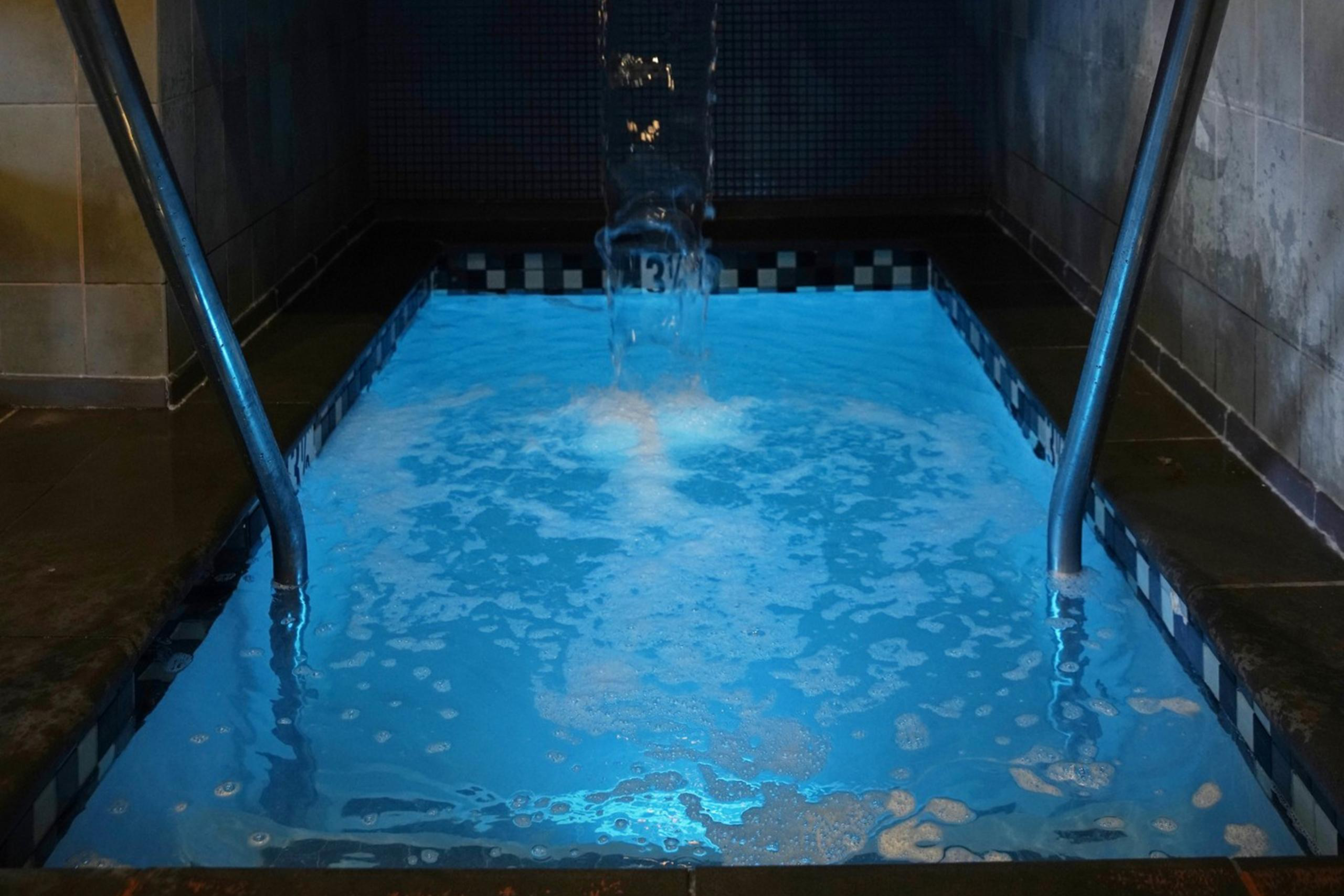 A small indoor pool with clear blue water features two handrails and a waterfall. Above, a painting depicts a person in a vintage bath setting. A clock shows 4:05.