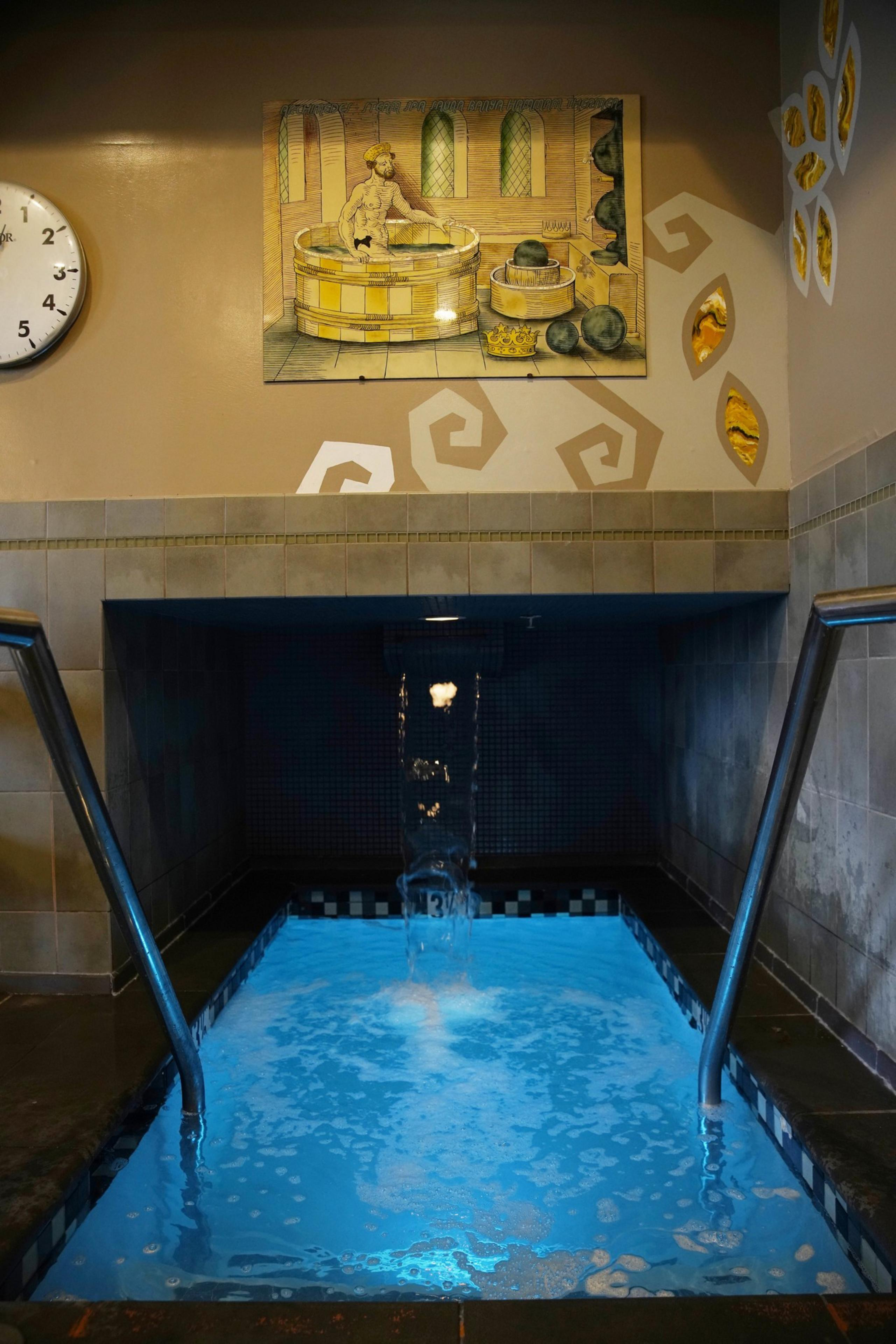 A small indoor pool with clear blue water features two handrails and a waterfall. Above, a painting depicts a person in a vintage bath setting. A clock shows 4:05.