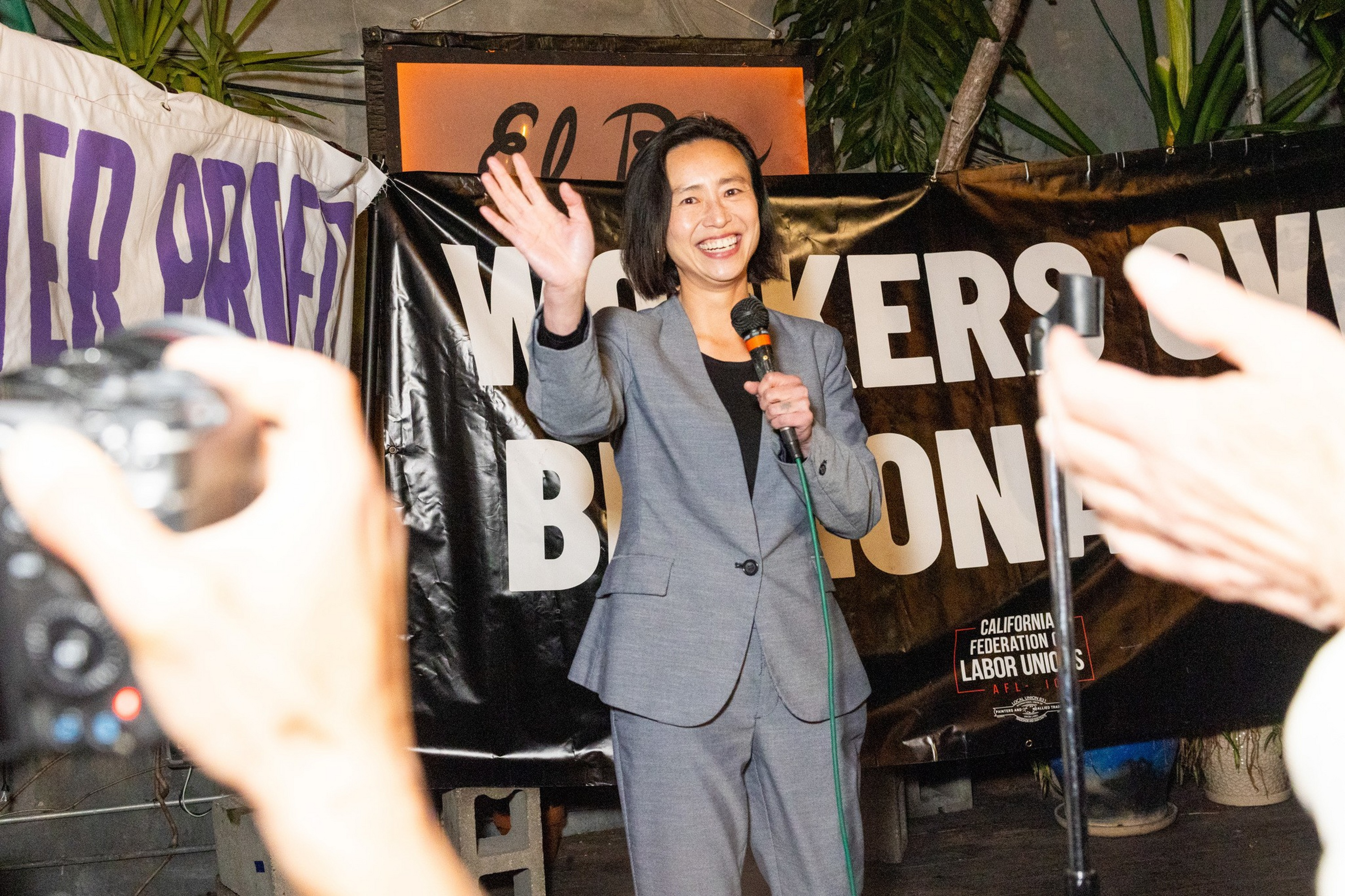 A woman in a gray suit smiles and waves while holding a microphone, standing in front of a banner that reads “Workers Over Billionaires.”