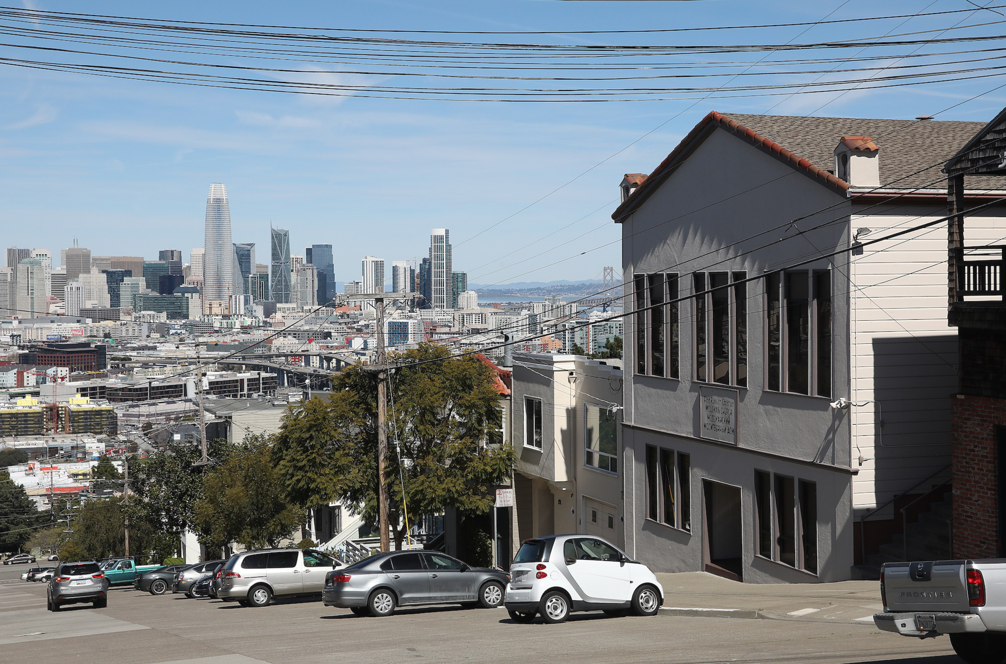 A steep city street with parked cars, power lines crisscrossing overhead, a large tree, and a city skyline in the background under a clear blue sky.