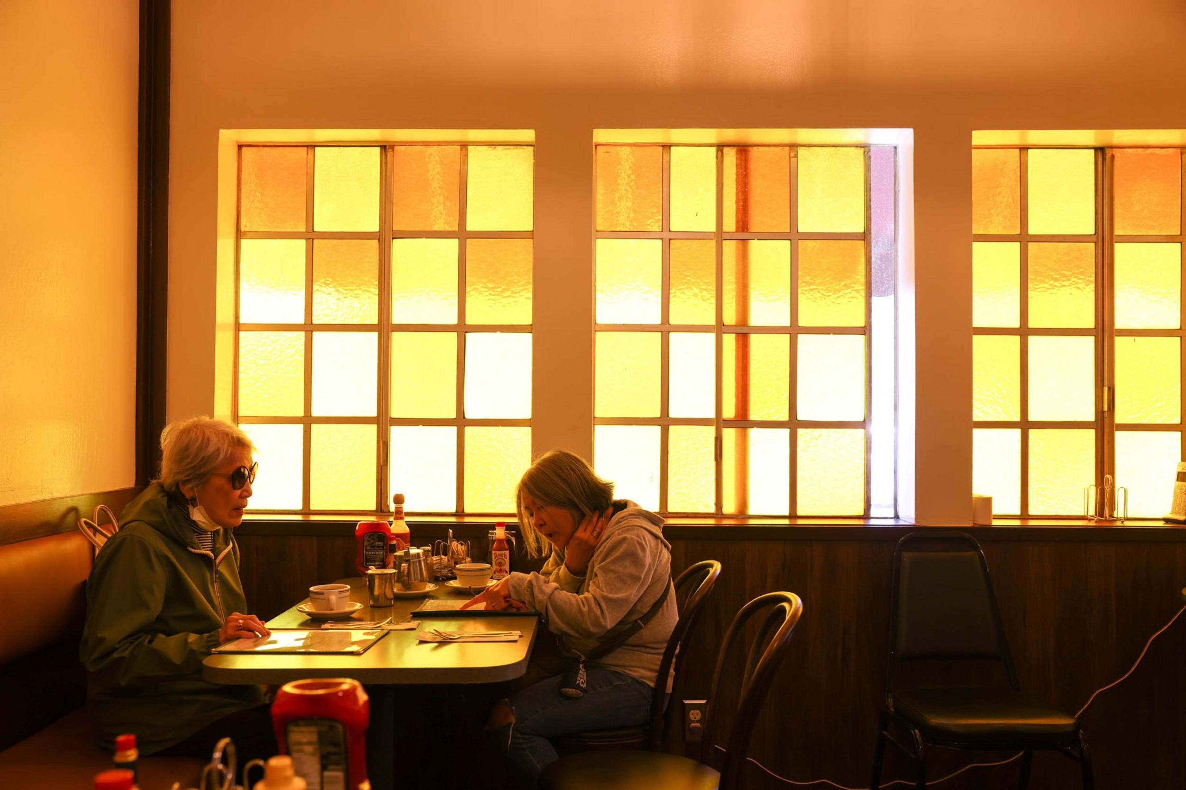 Two women are sitting at a table in a cafe with orange-tinted windows. The table has cups and condiments. One woman wears sunglasses, the other reads a menu.