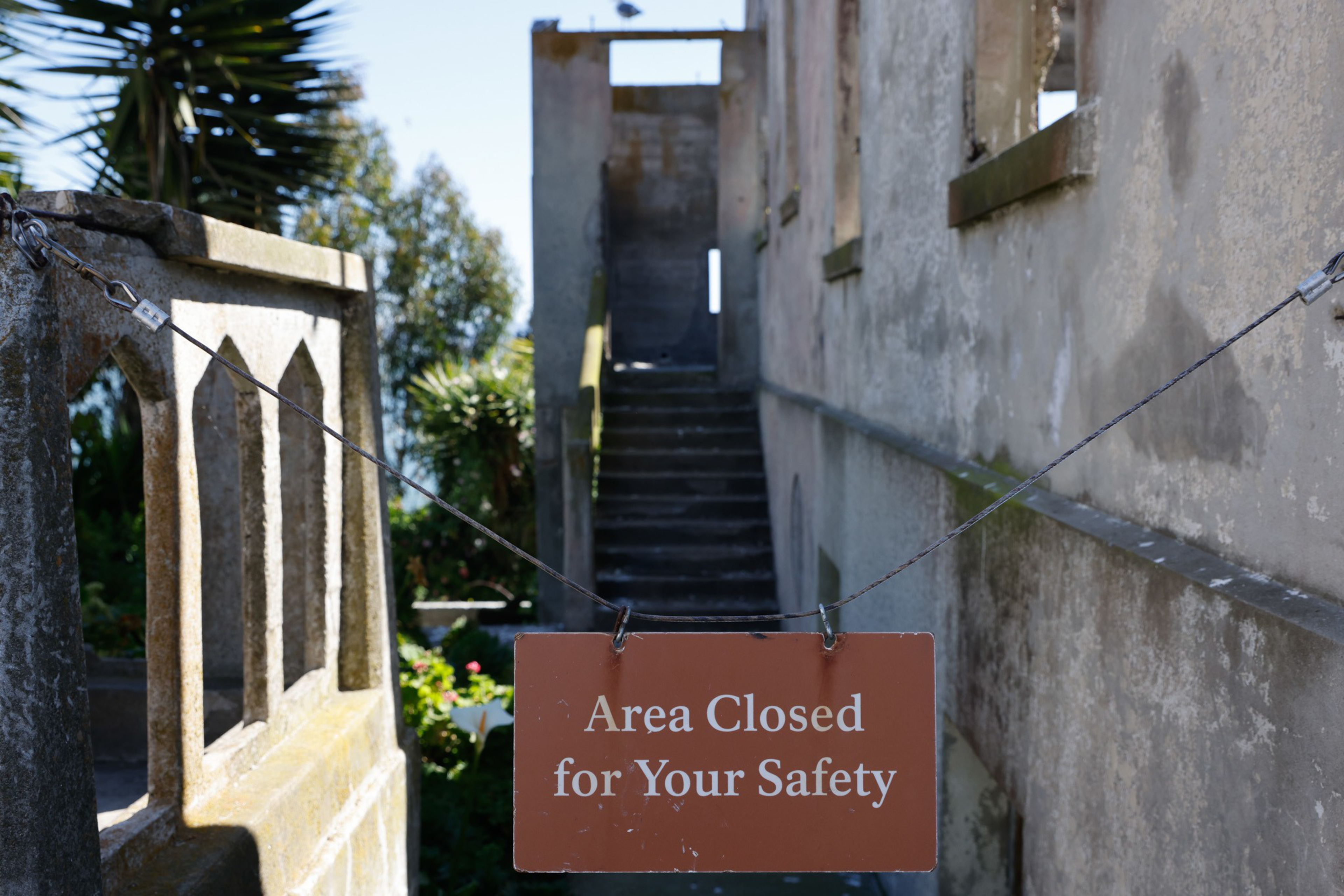 A weathered stone staircase is blocked by a chain with a sign saying &quot;Area Closed for Your Safety.&quot; Surrounding greenery contrasts with the decaying structure.