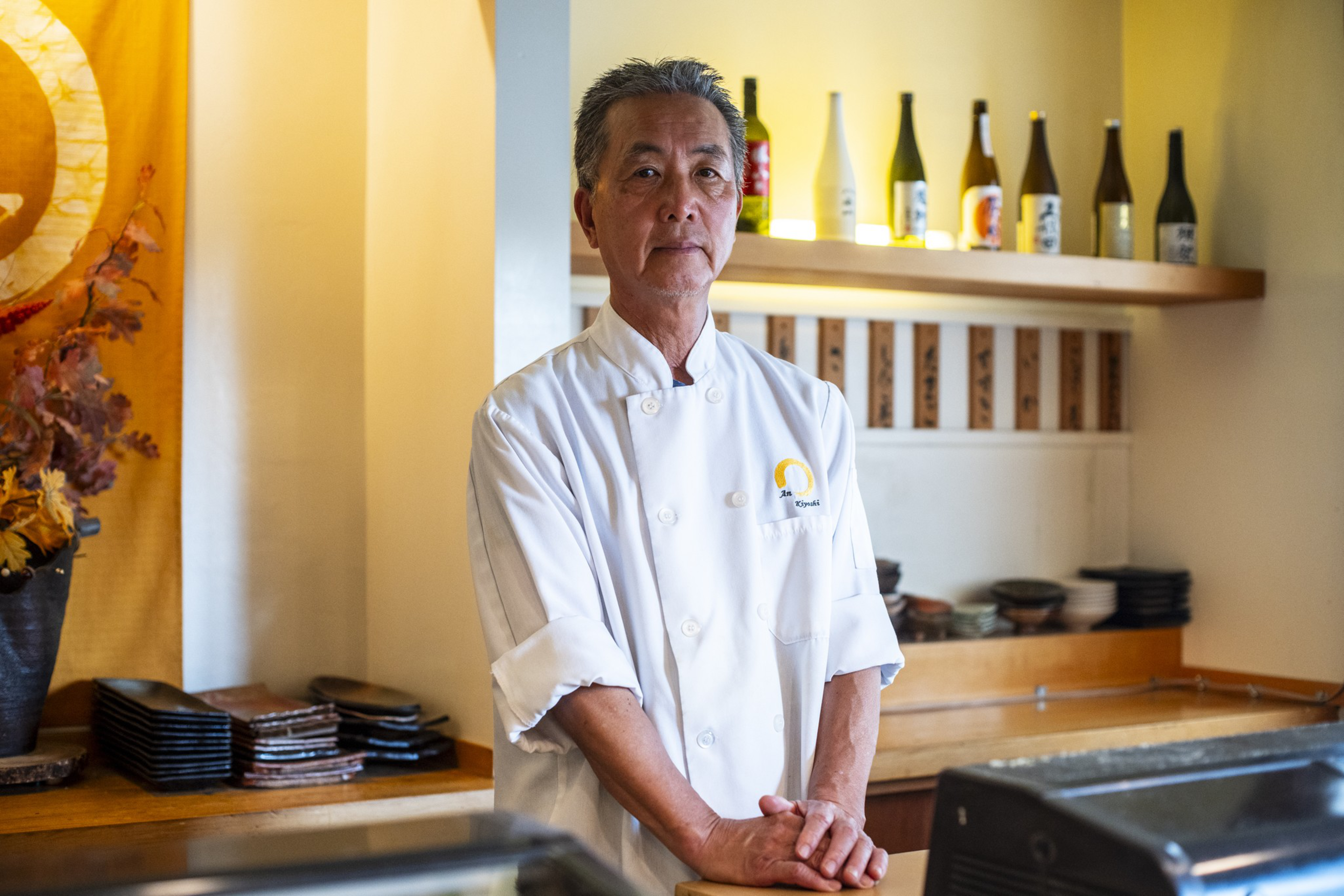 A man in a white chef's coat stands behind a wooden counter in a restaurant with bottles on a lit shelf and stacked plates in the background.