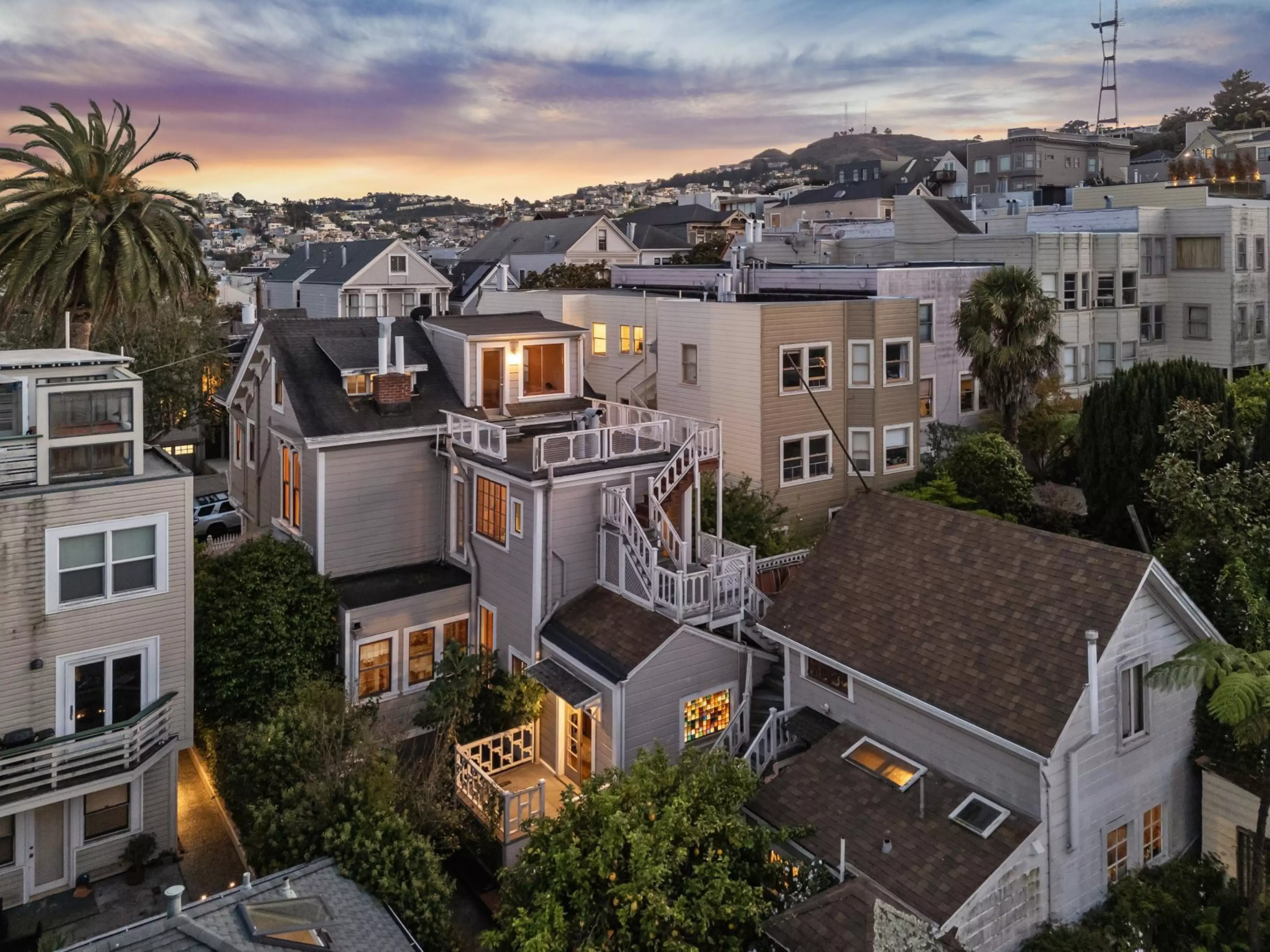 A neighborhood of closely packed houses with lit windows at dusk, palm trees, and hills in the background under a colorful sunset sky.