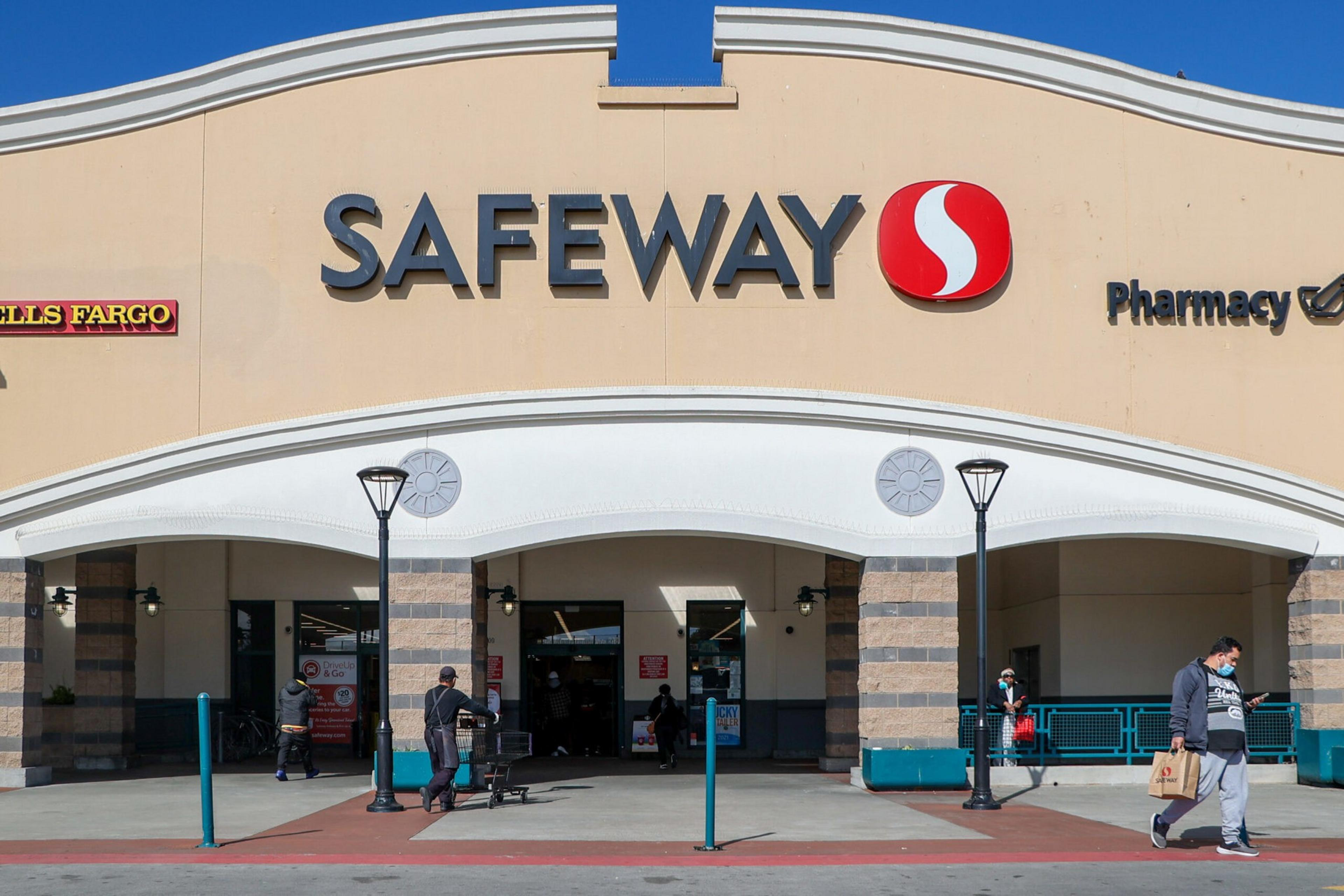 A Safeway store entrance with people wearing masks, a person pushing a shopping cart, and a wall sign for Wells Fargo and pharmacy services.