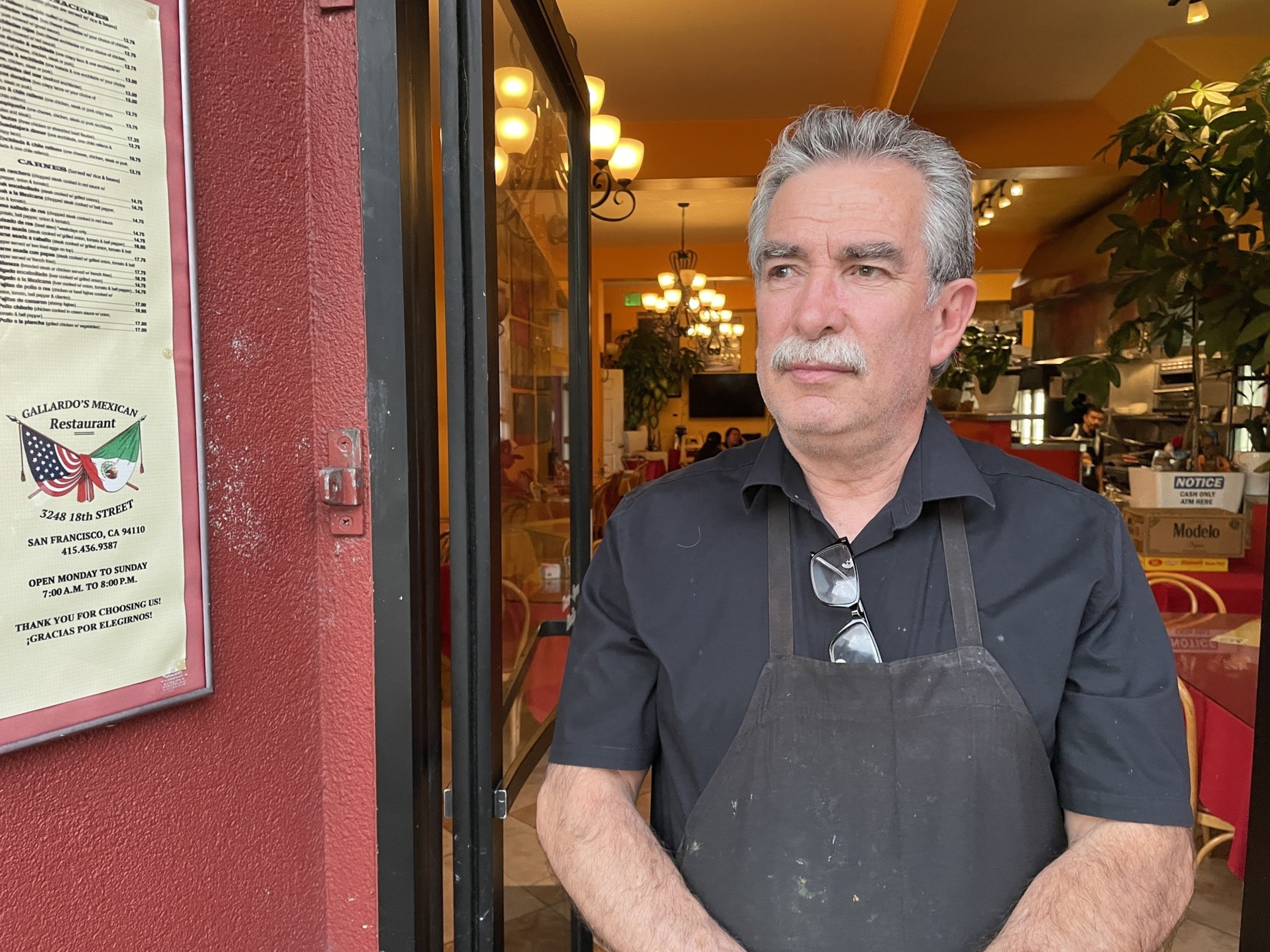 A man with gray hair and a mustache stands at the entrance of a Mexican restaurant, wearing a black shirt and apron. A menu is displayed on the wall beside him.