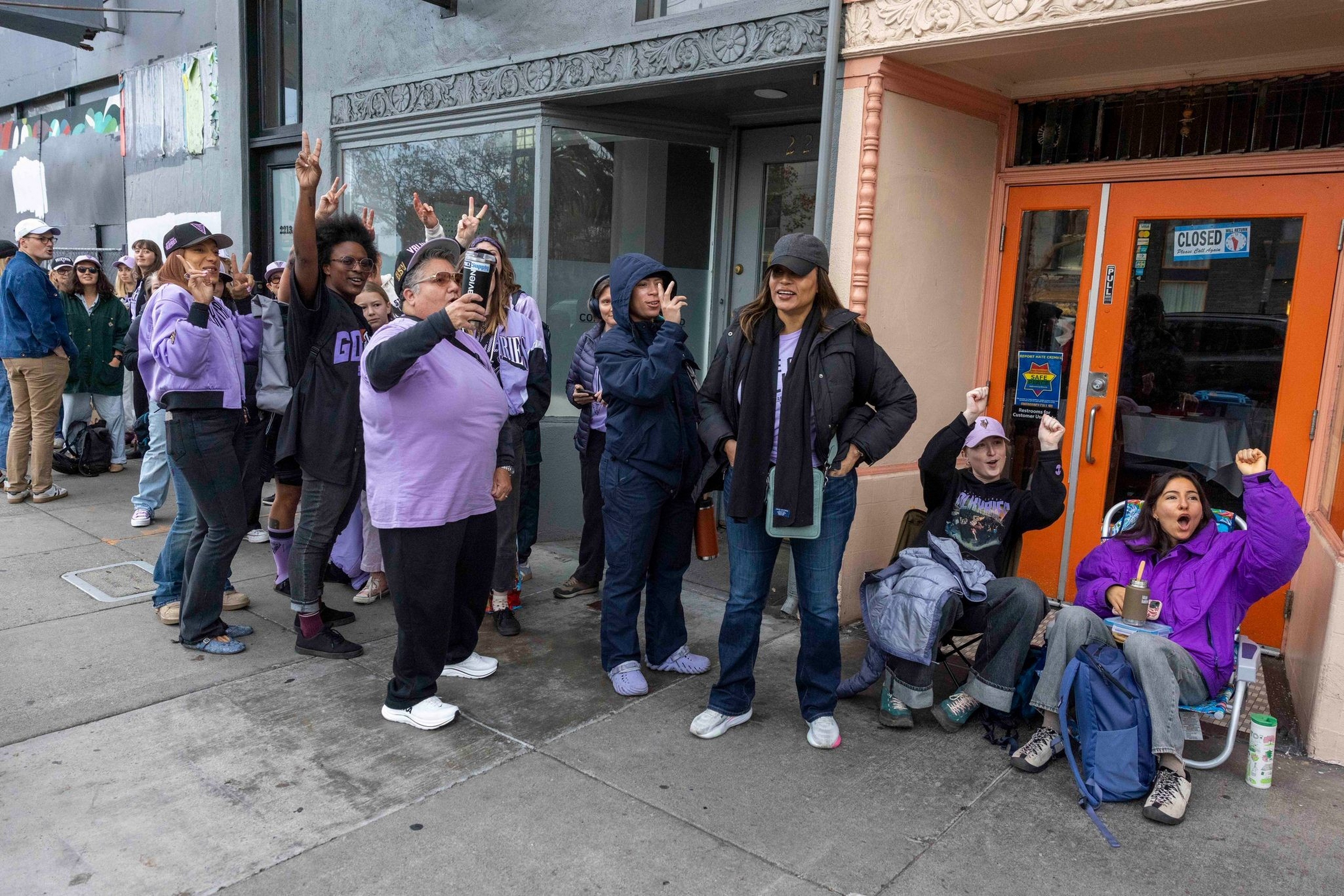 A diverse group of people, many wearing purple, stand and sit excitedly outside a closed store with an orange door.