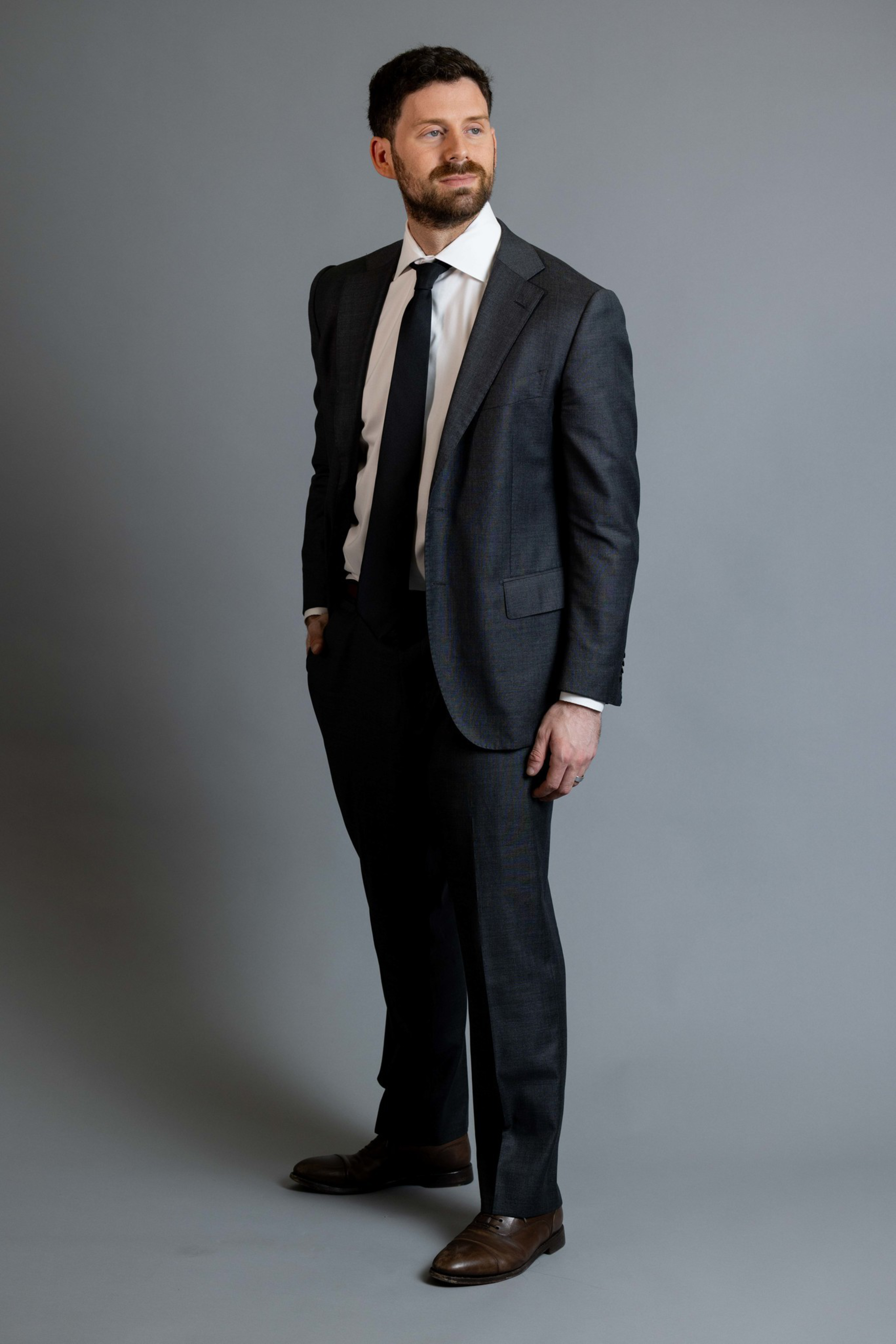 A man with short dark hair and a beard wears a dark suit, white shirt, black tie, and brown shoes, standing with one hand in his pocket.