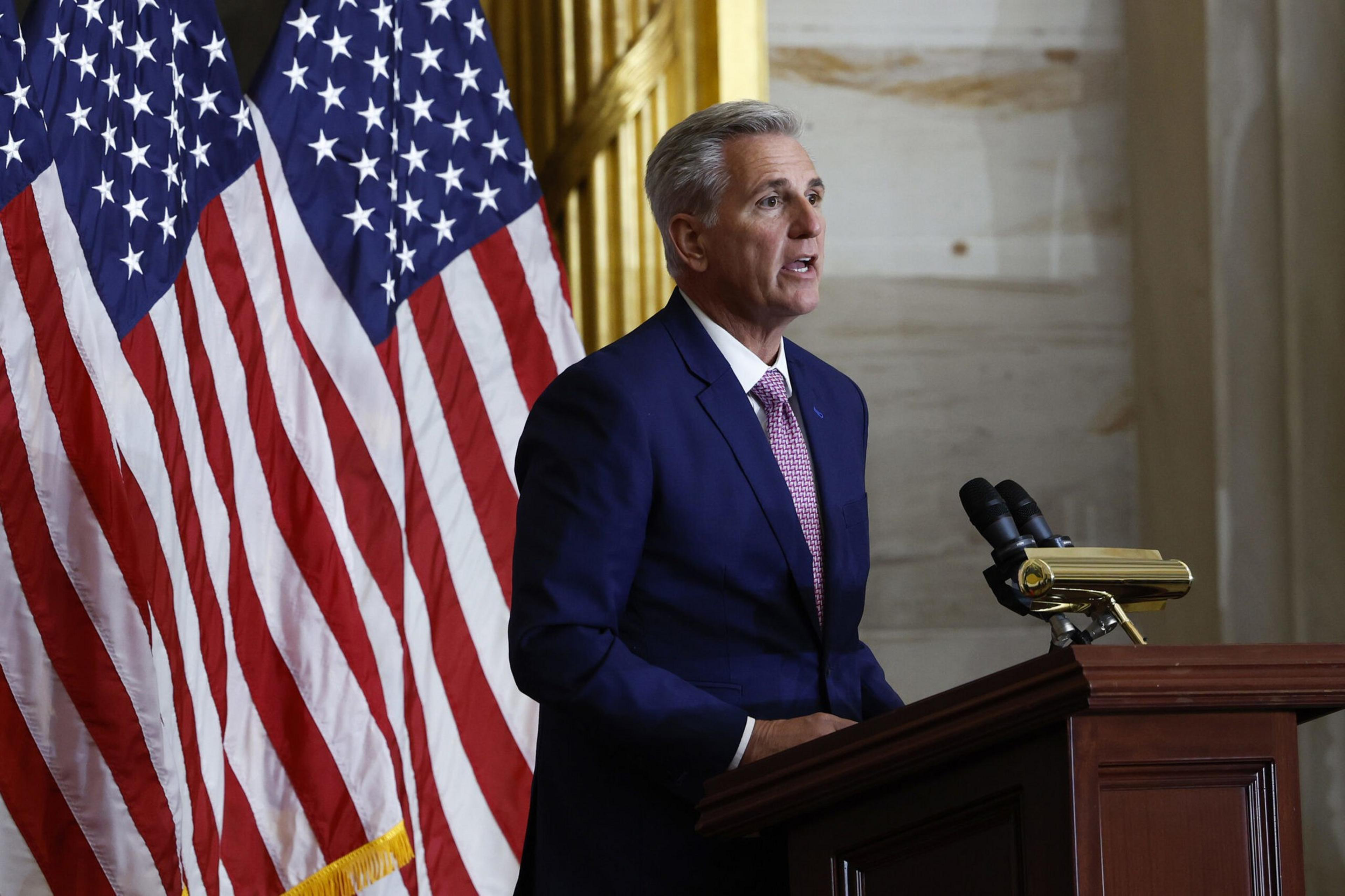 A man in a blue suit and pink tie speaks at a wooden podium with microphones, standing in front of two American flags.