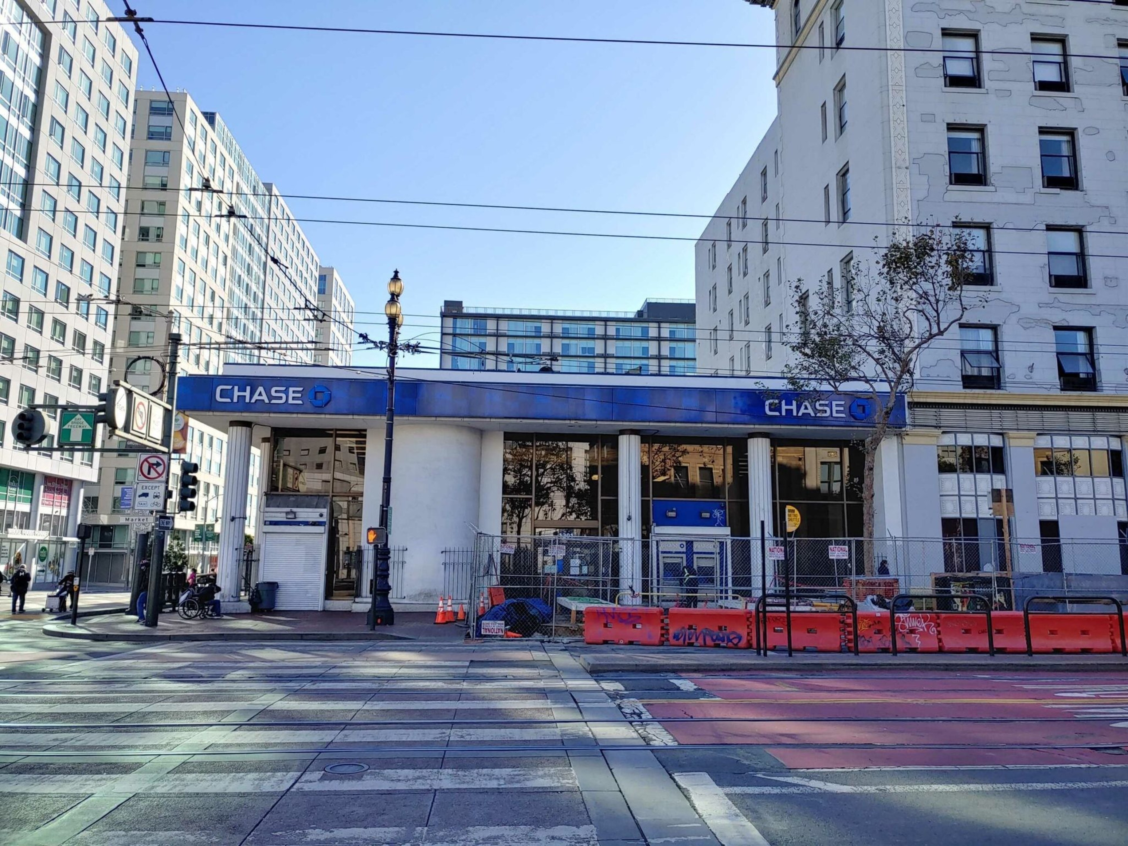 A Chase bank branch is behind orange construction barriers on a city street corner, with a crosswalk and traffic lights visible.