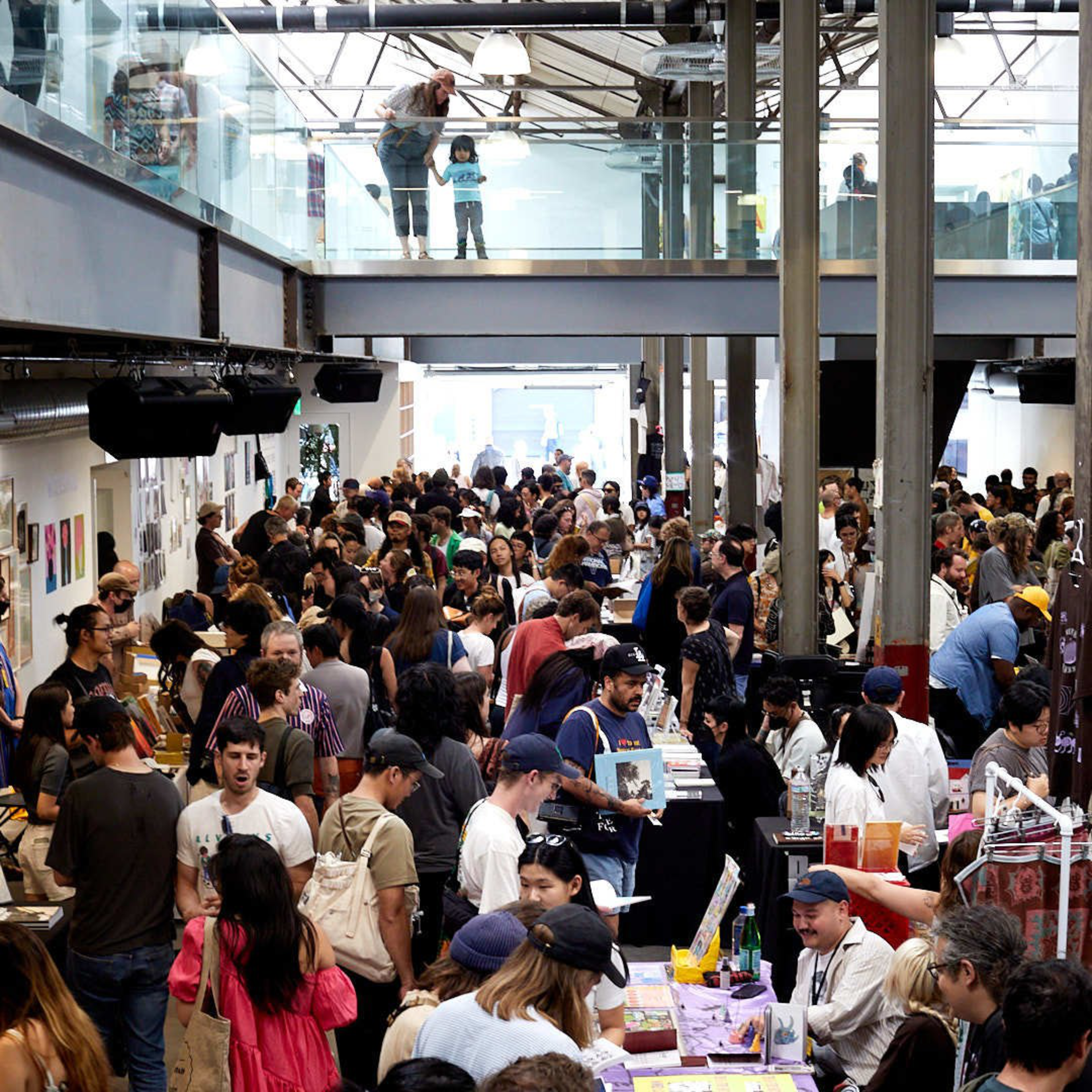 A busy indoor market or fair with crowded tables displaying various items. People are interacting, and a glass upper level shows a few more observers.
