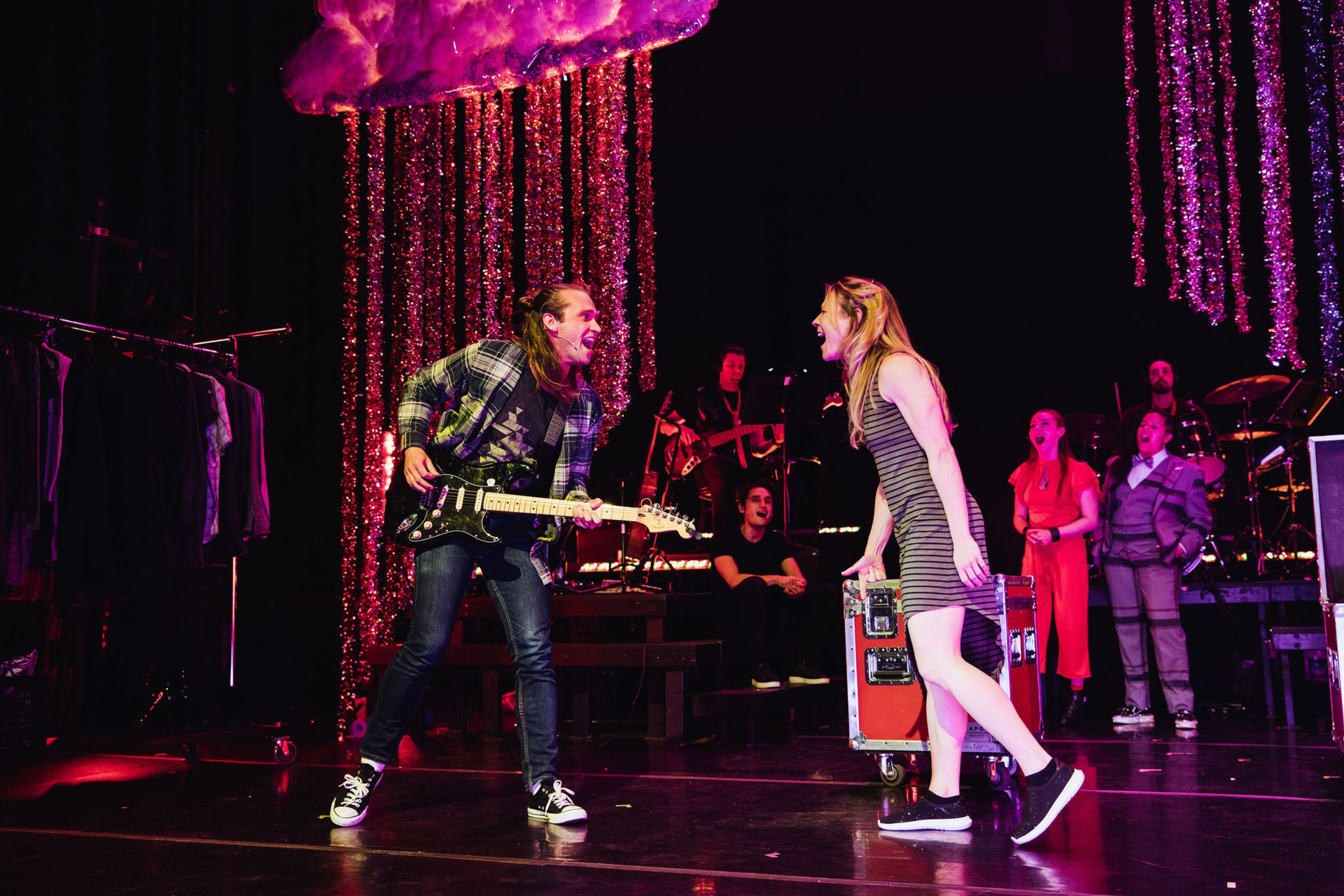 A man plays electric guitar passionately while a woman in a striped dress and sneakers leans forward, singing or shouting at him on stage.