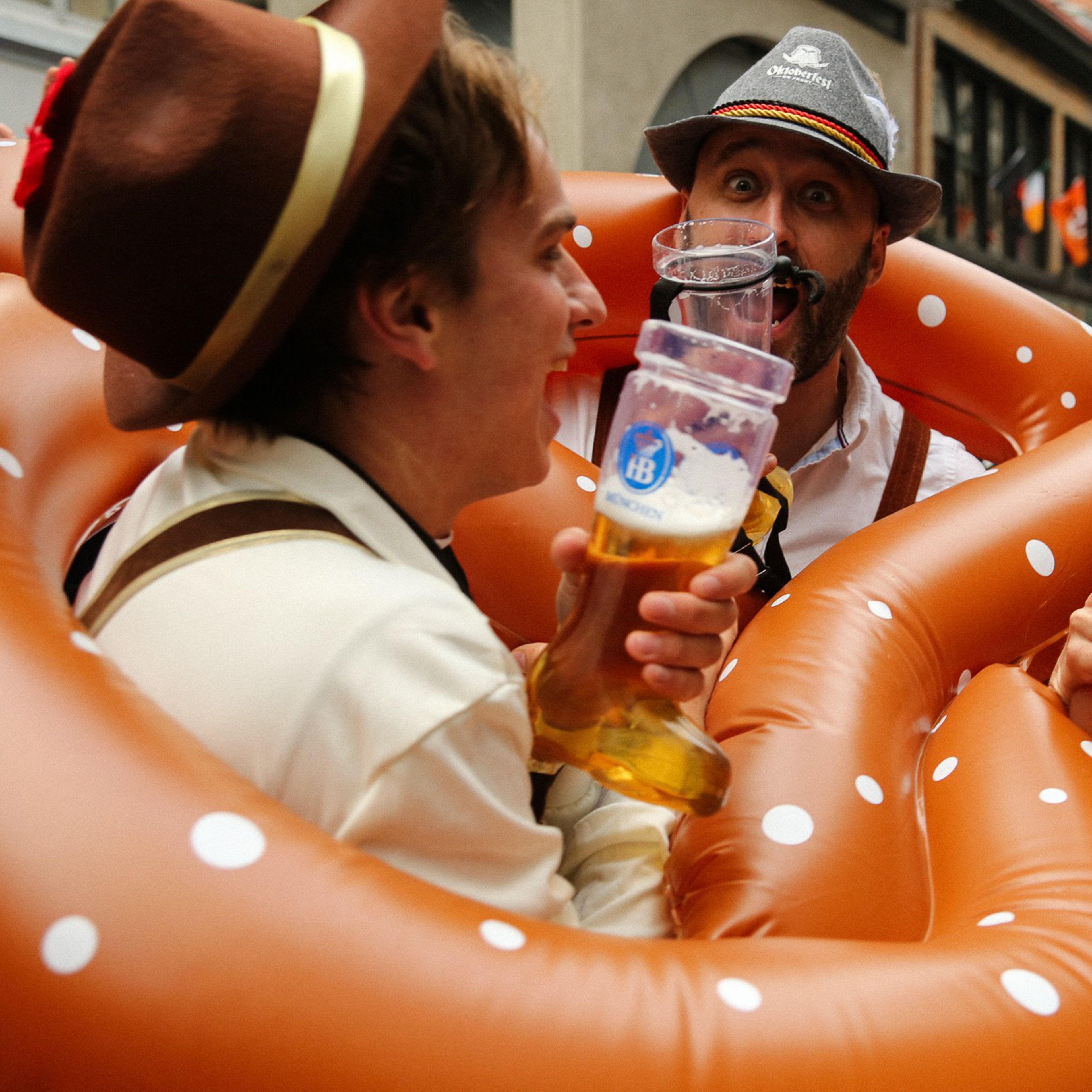 Three people in festive attire drink from large beer glasses, surrounded by inflatable pretzels, celebrating together in an outdoor setting.