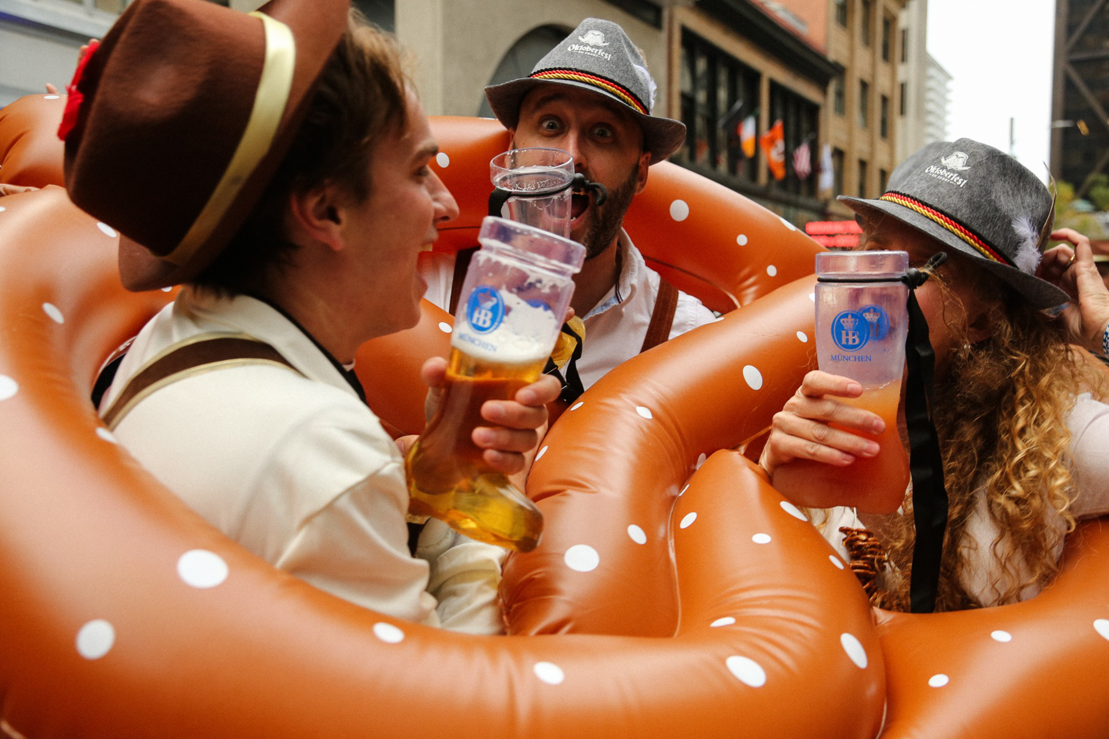 Three people in festive attire drink from large beer glasses, surrounded by inflatable pretzels, celebrating together in an outdoor setting.