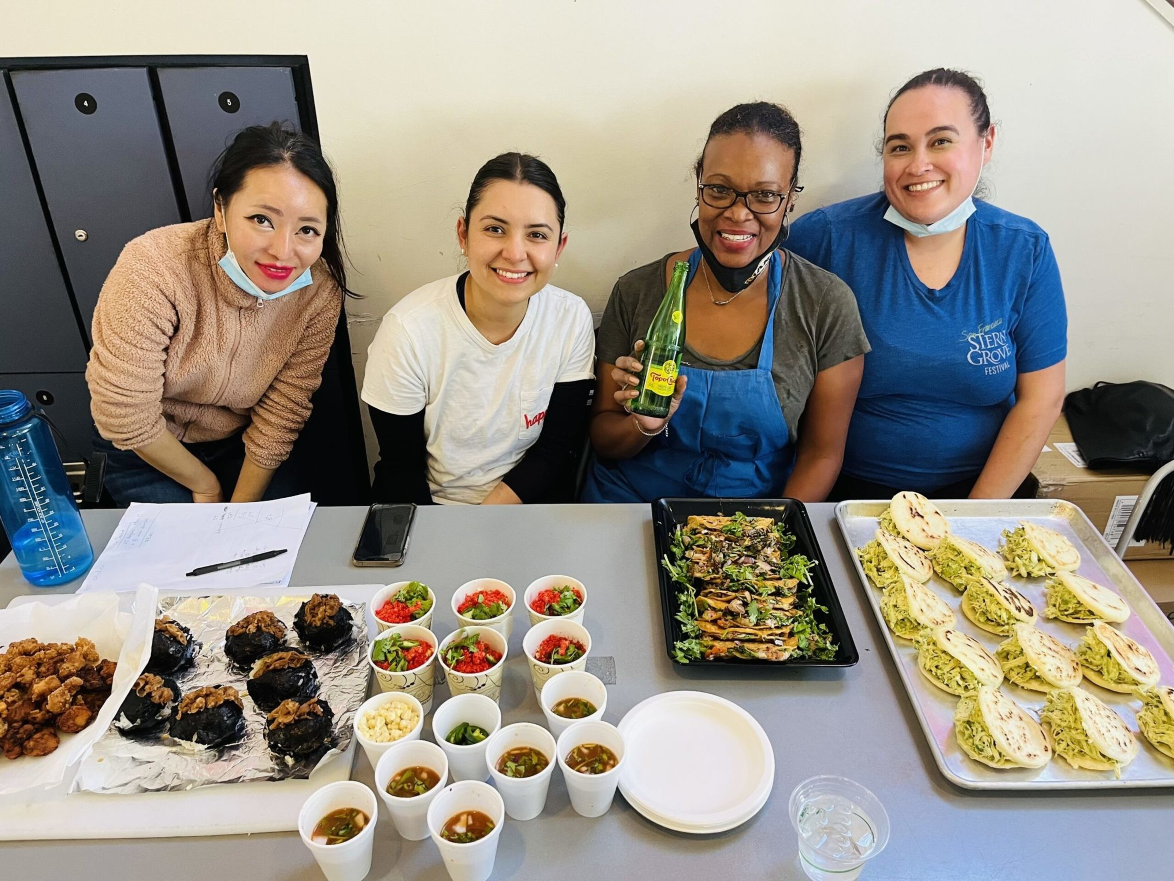 Four women smile behind a table filled with various foods like fried bites, cups of soups, salads, grilled vegetables, and sandwiches, one holding a green bottle.