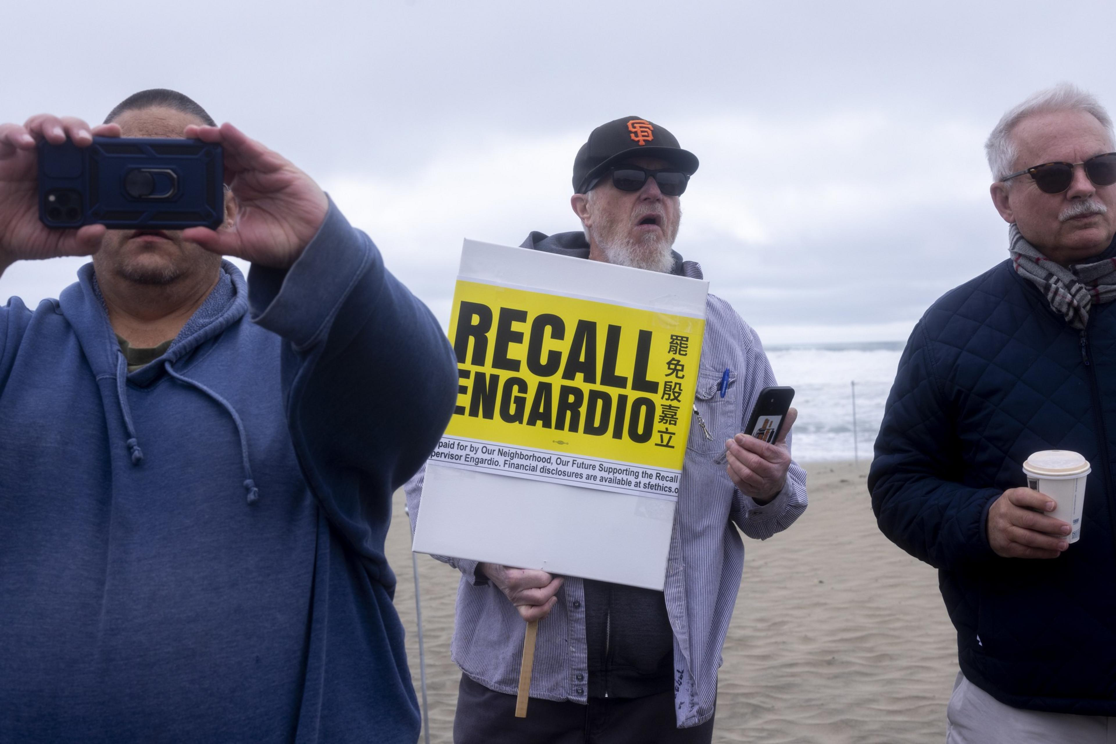 Three men stand on a beach, one holding a "RECALL ENGARDIO" sign in yellow and black, another taking a photo, and the third holding a coffee cup.