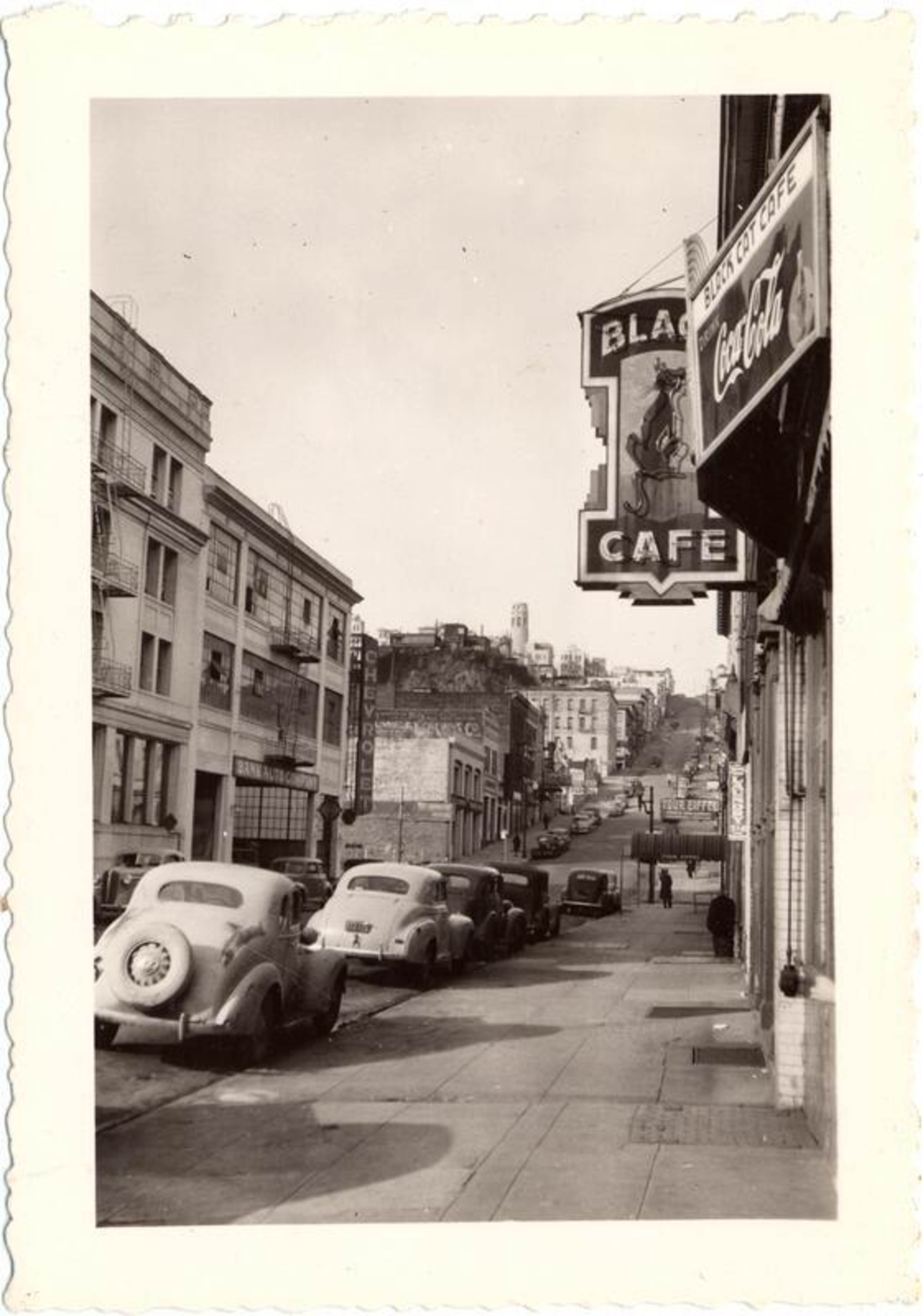 A steep city street lined with vintage cars is flanked by buildings, with signs for a café and Coca-Cola visible on the right side.