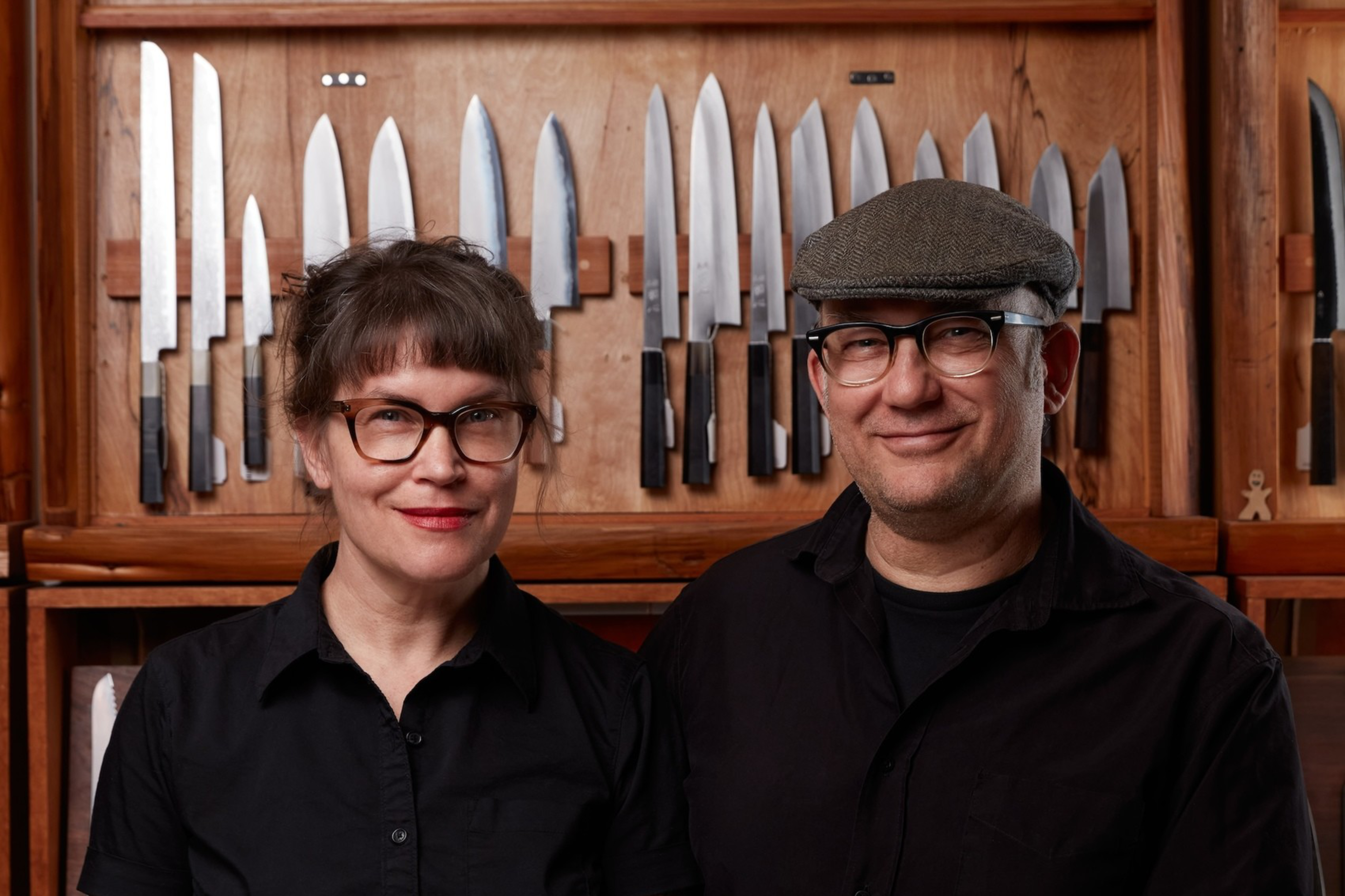 Two people, a women wearing glasses and a black shirt and a man wearing glasses and a hat also in a black shirt, stand behind a counter displaying knives.