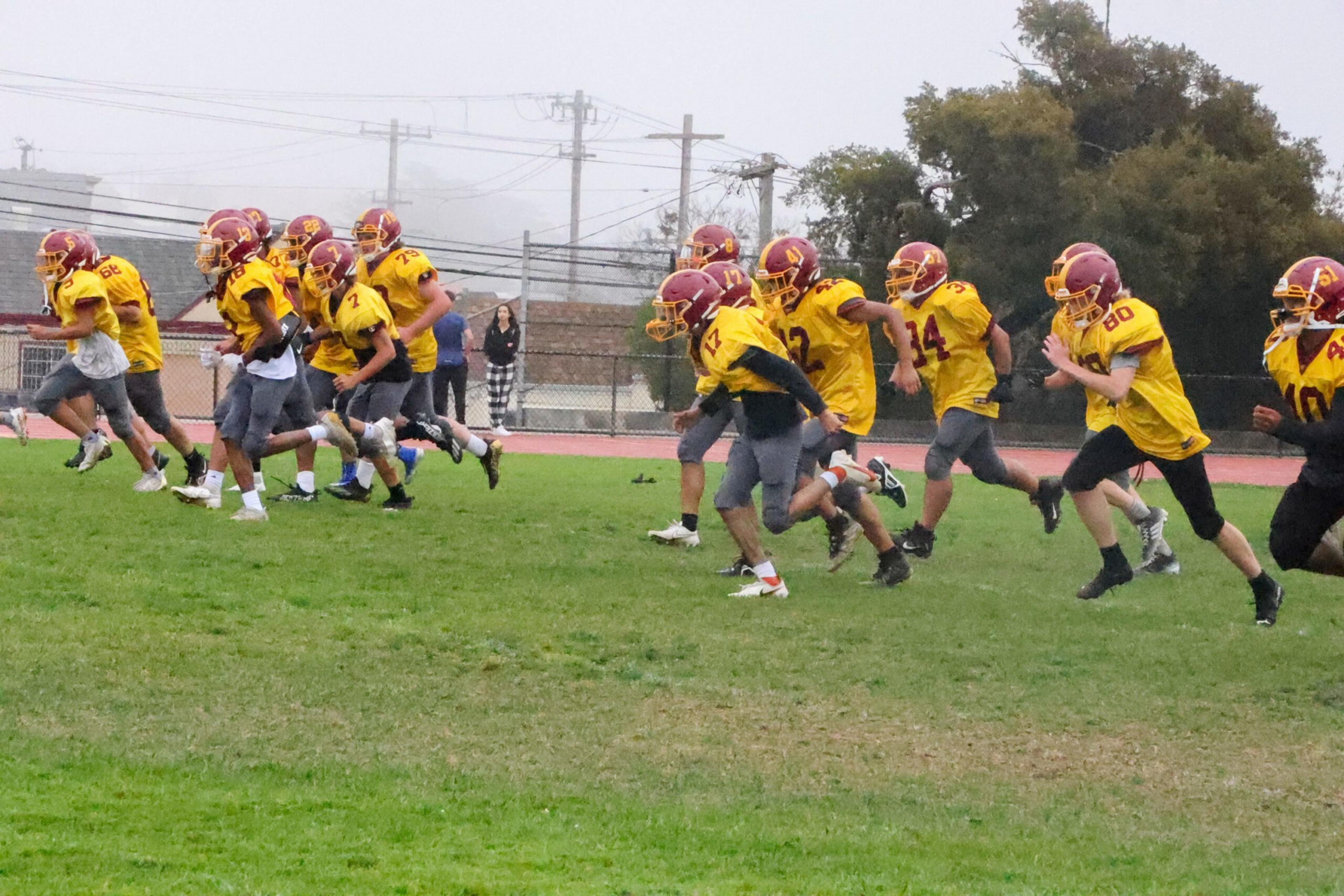 A group of youth football players in yellow jerseys and red helmets are running and practicing on a grassy field.