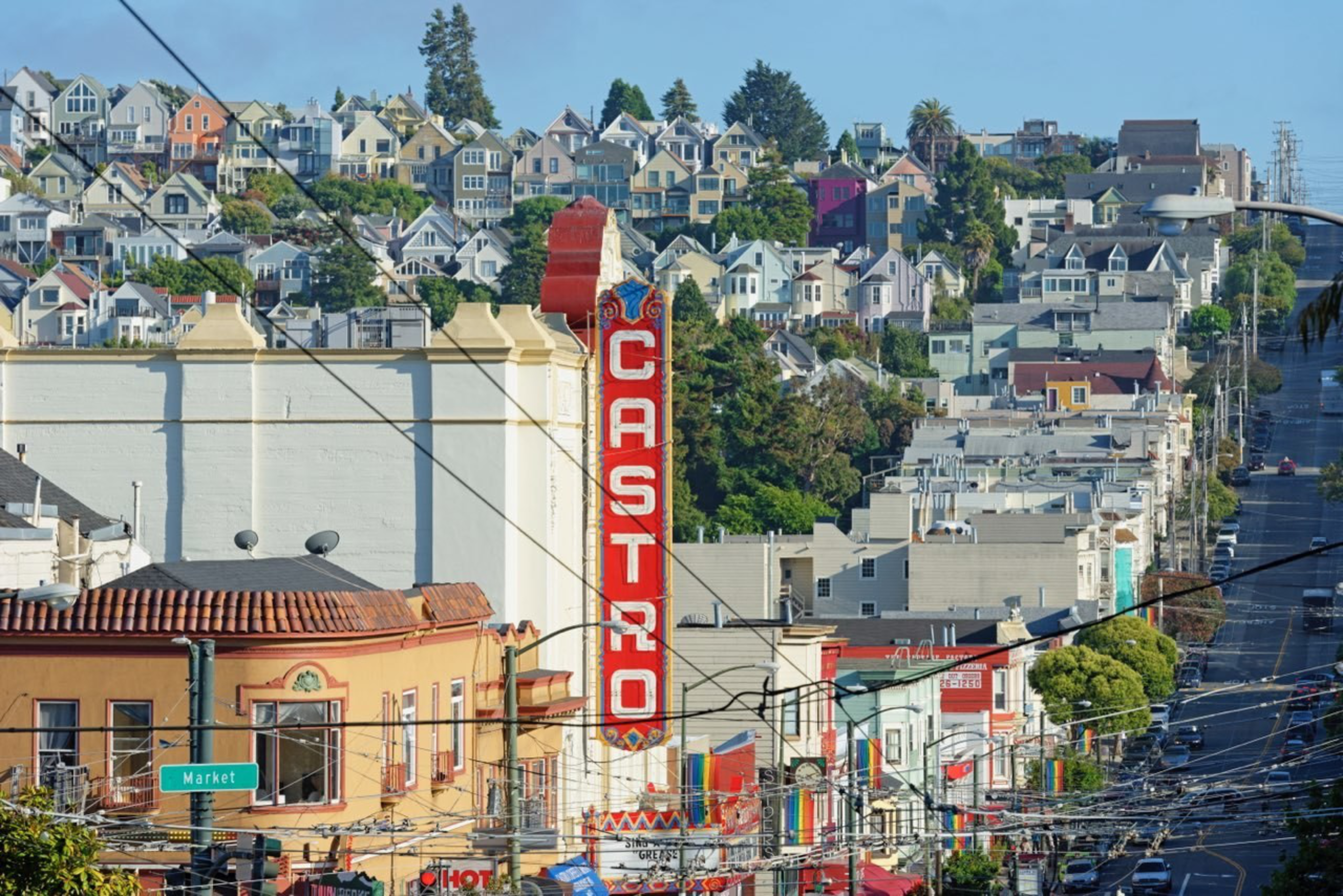 A vibrant urban street lined with colorful houses on a hillside, featuring a tall red "CASTRO" sign and numerous power lines crossing overhead.