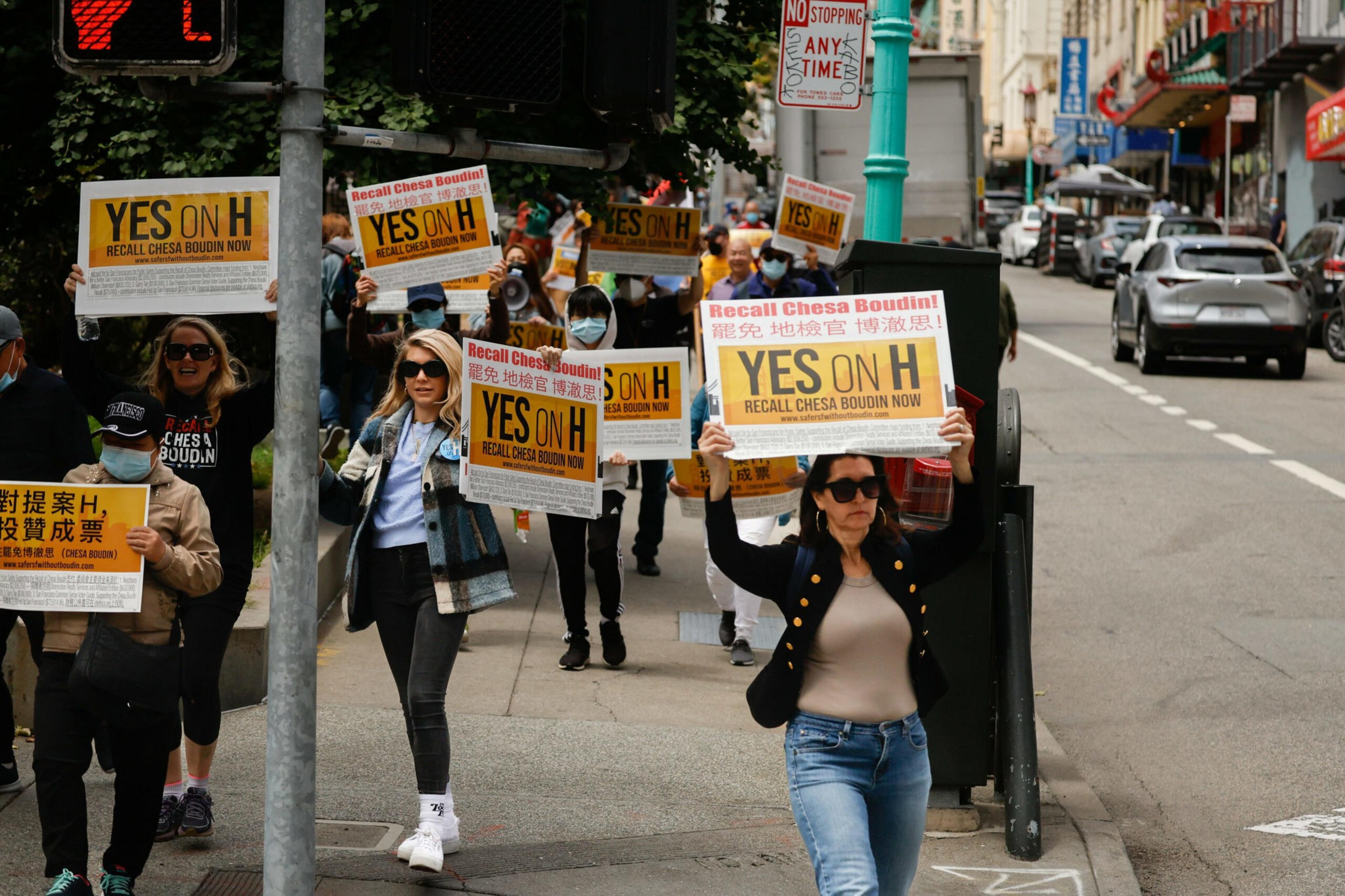 People march on a city sidewalk holding signs that say "YES ON H" and "Recall Chesa Boudin Now" in English and Chinese.