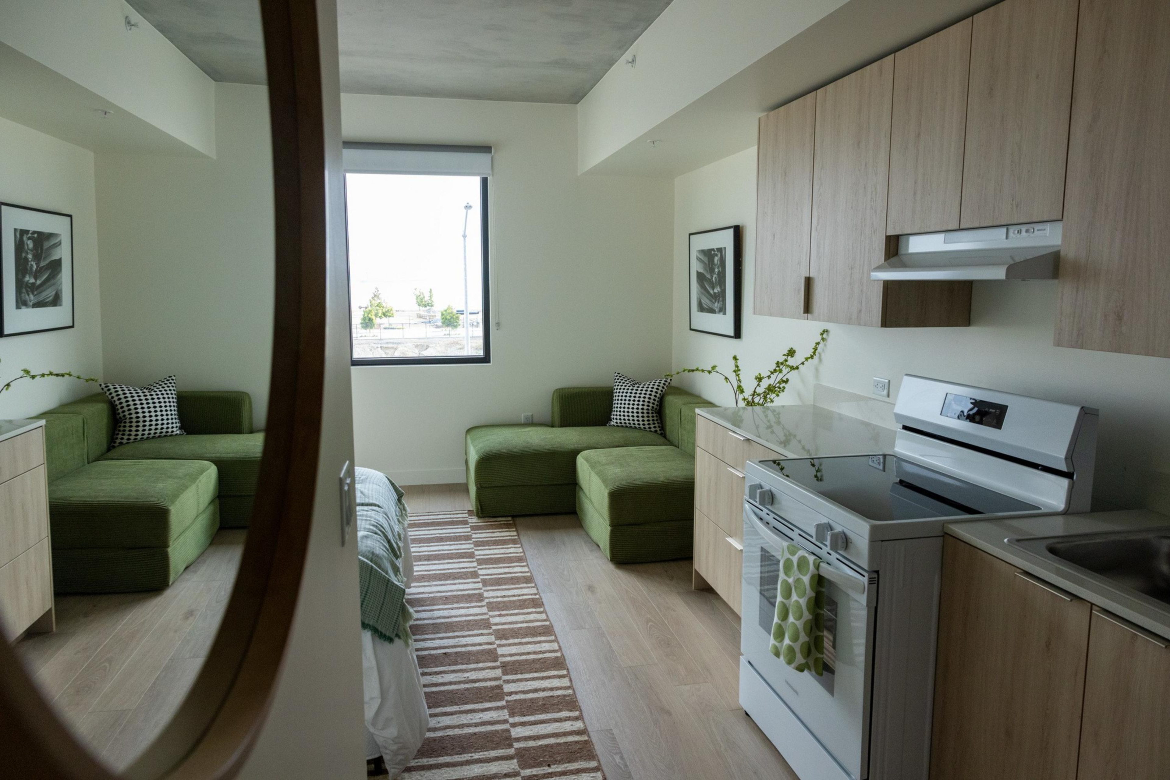 A small kitchen area with light wood cabinets, a white stove, and a green sofa with pillows, reflected in a large round mirror.