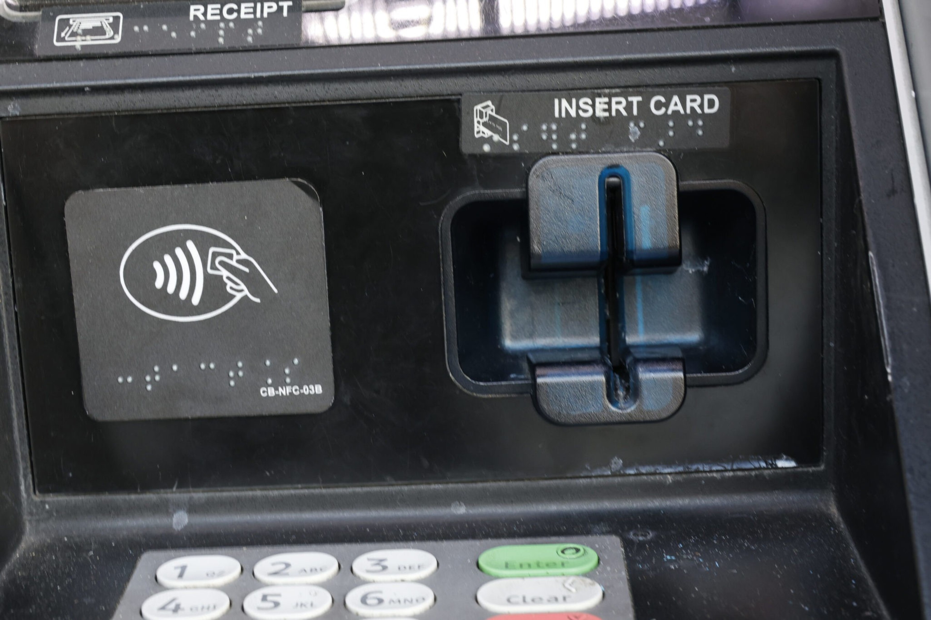 A payment terminal with a card insertion slot, contactless payment icon, Braille markings, and a numeric keypad with green "Enter" and red "Clear" buttons.