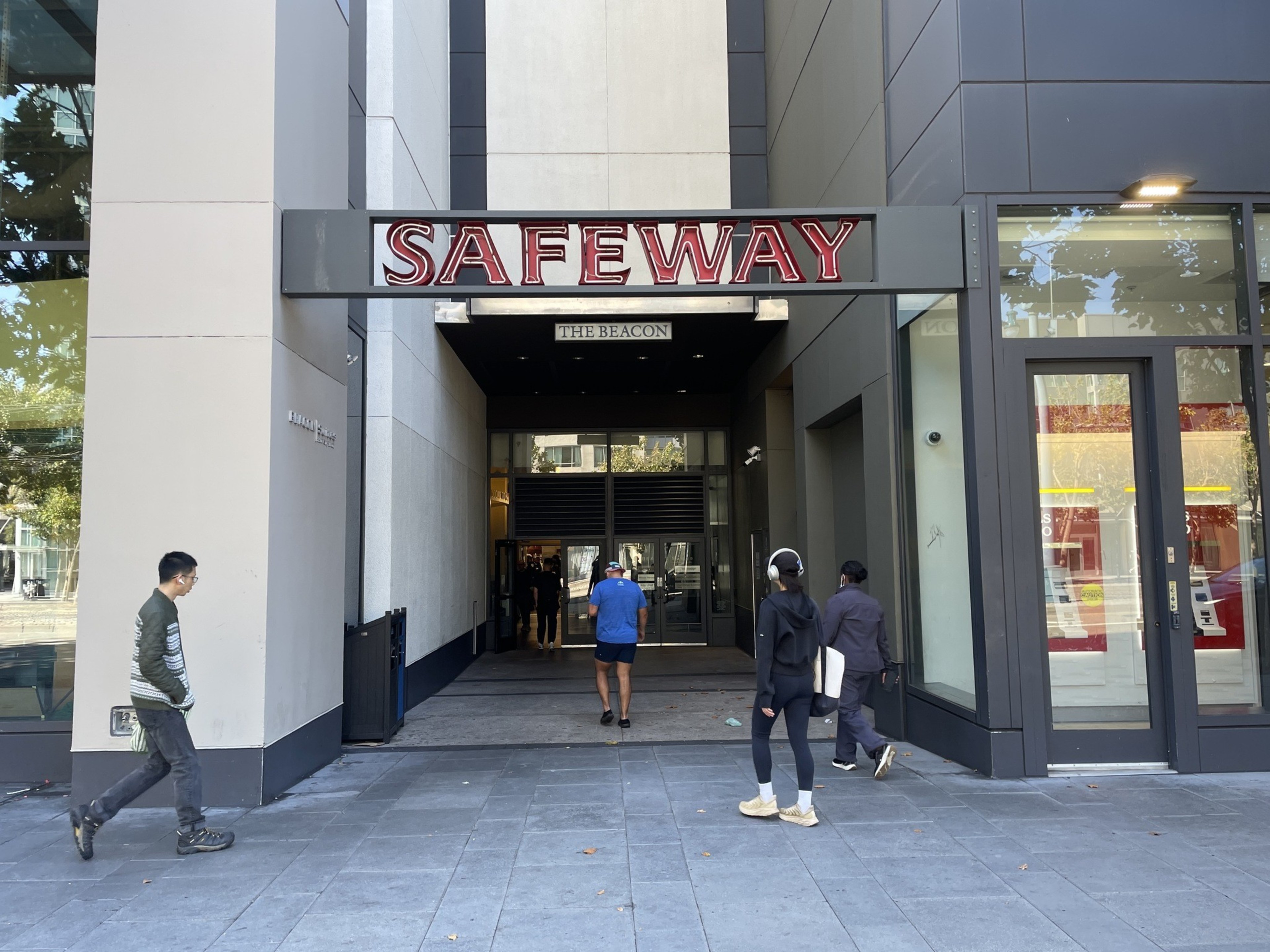 Several people walk near the entrance of a Safeway store called "The Beacon," located between tall modern buildings with reflective glass and gray walls.