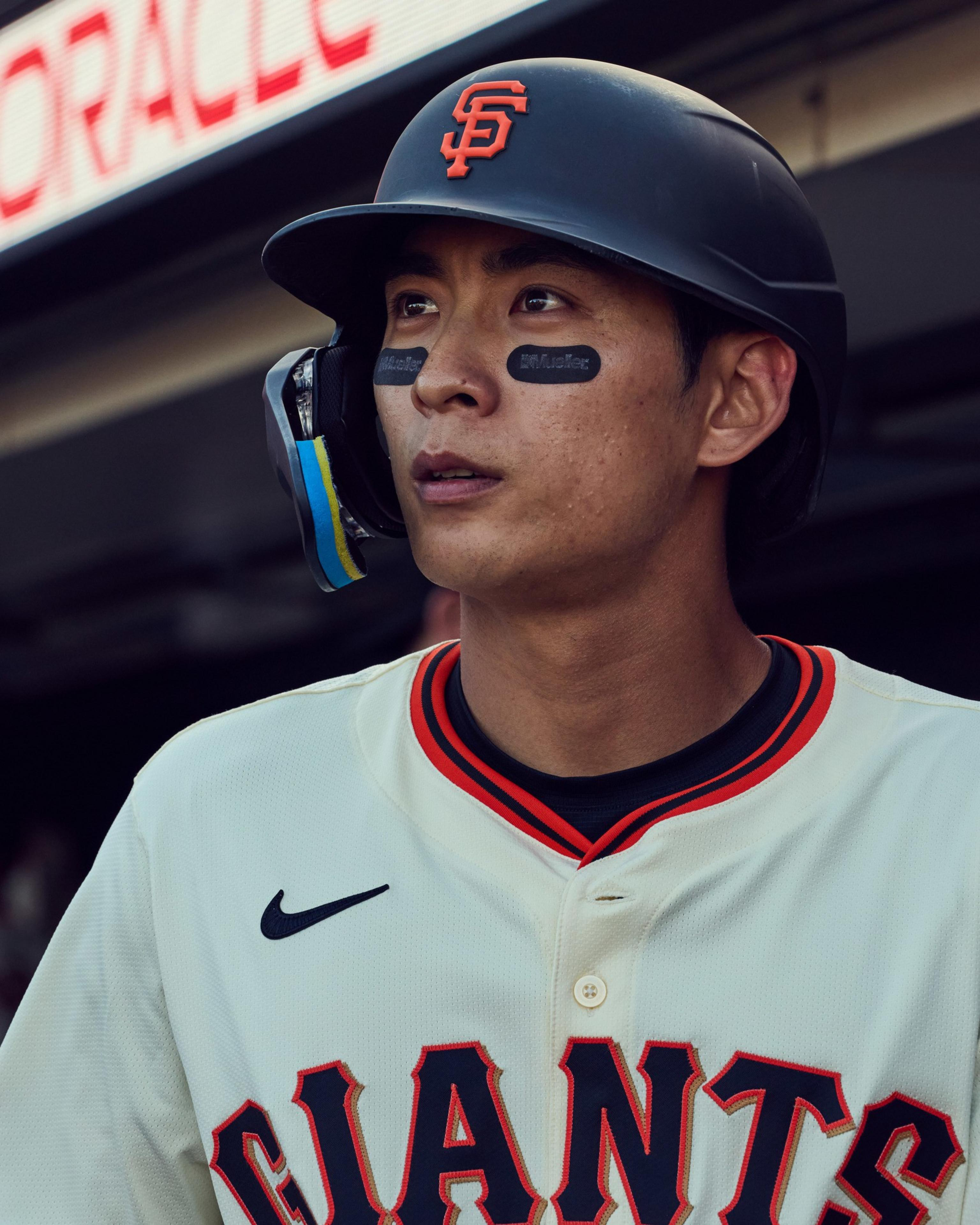 A baseball player wearing a helmet and a Giants jersey, with eye black under his eyes, looks focused in a dugout setting.