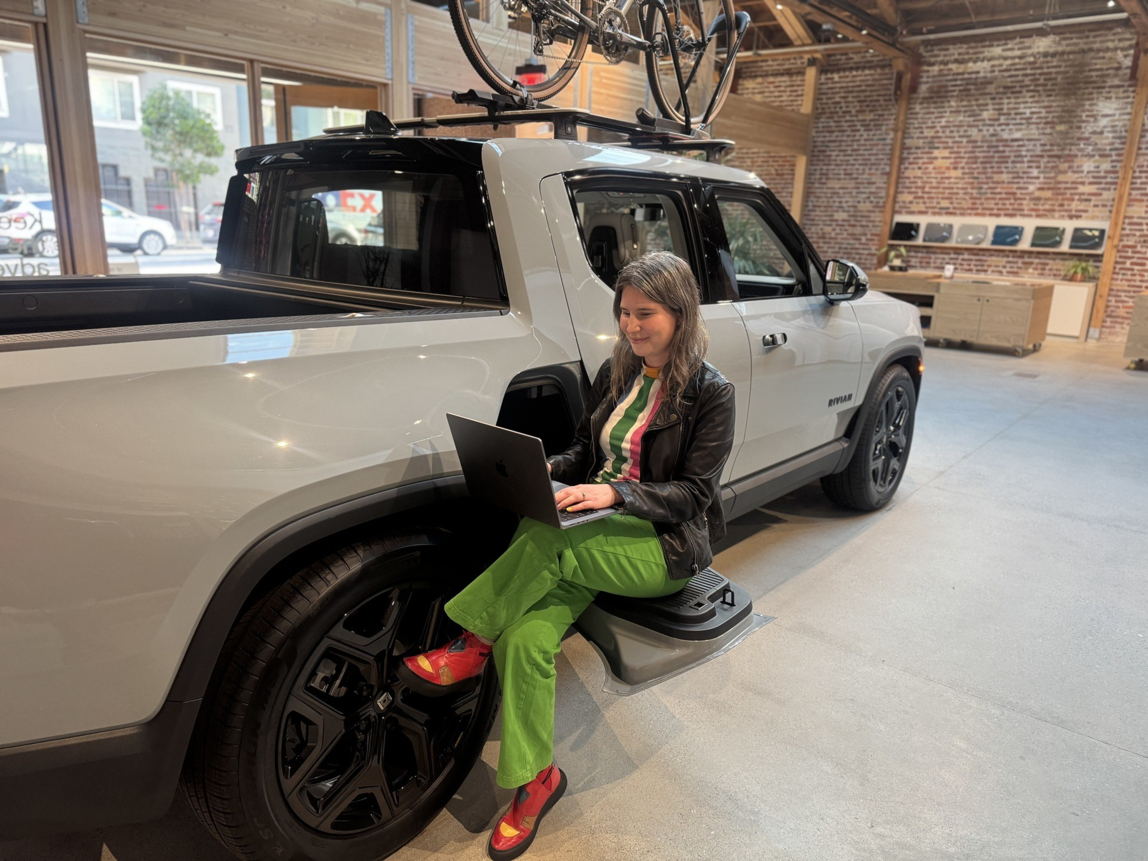 A woman in colorful attire sits on a truck's sidestep, using a laptop. The truck is indoors with bicycles on its roof and a brick wall in the background.