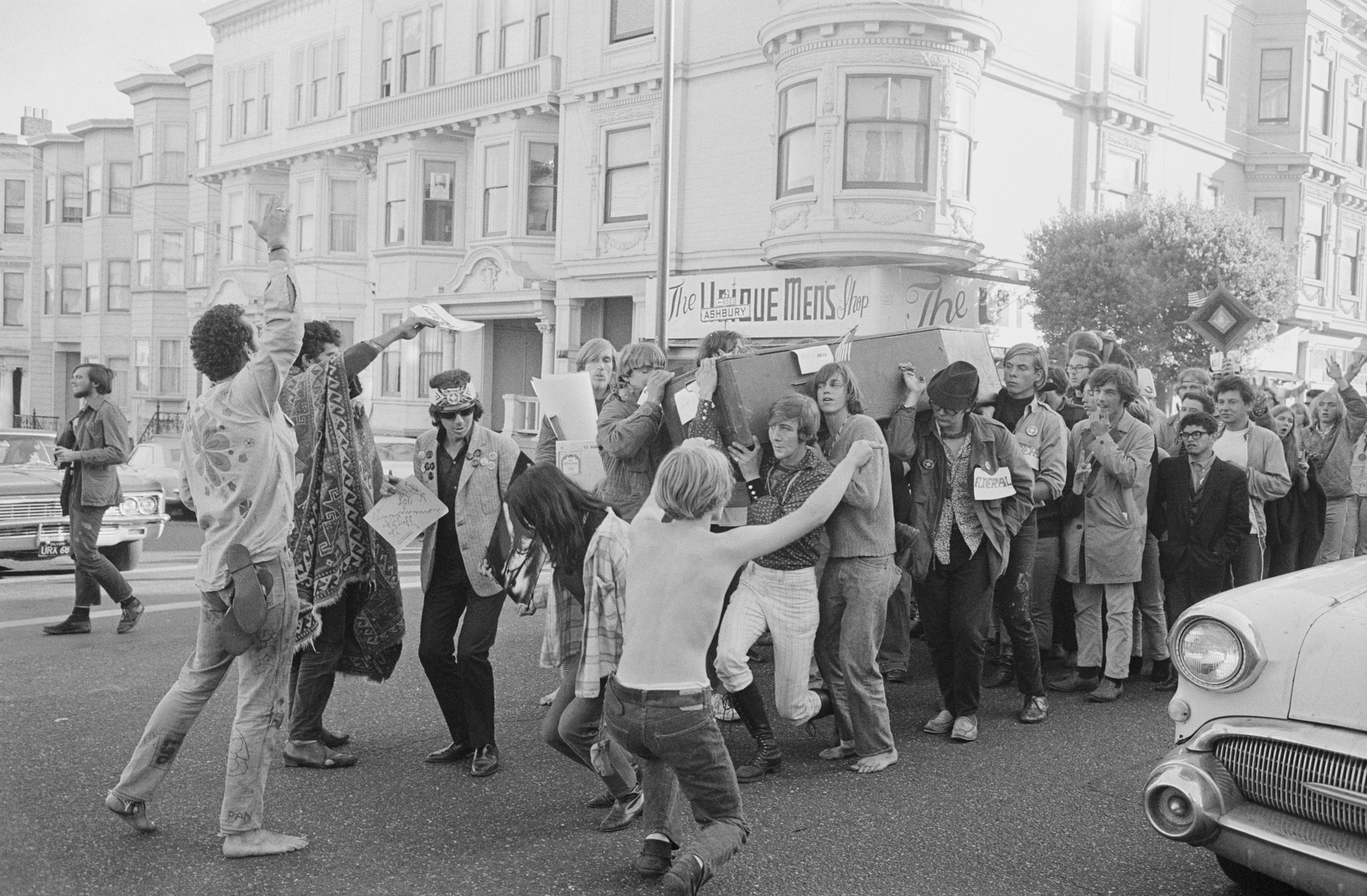 A group of people carry a coffin down a city street, surrounded by others dancing, holding signs, and raising hands in protest or celebration.