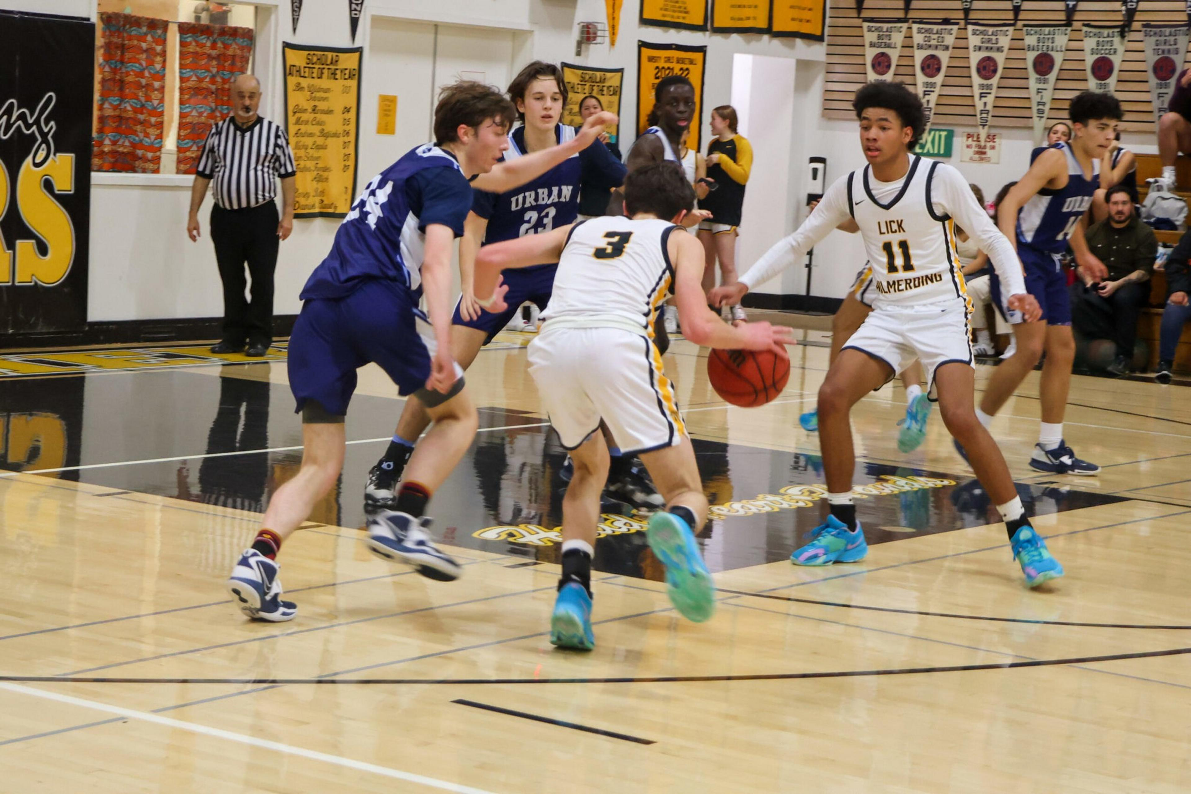 Two basketball players in white jerseys are on offense, with one dribbling the ball, while multiple players in blue jerseys are defending on the gym court.