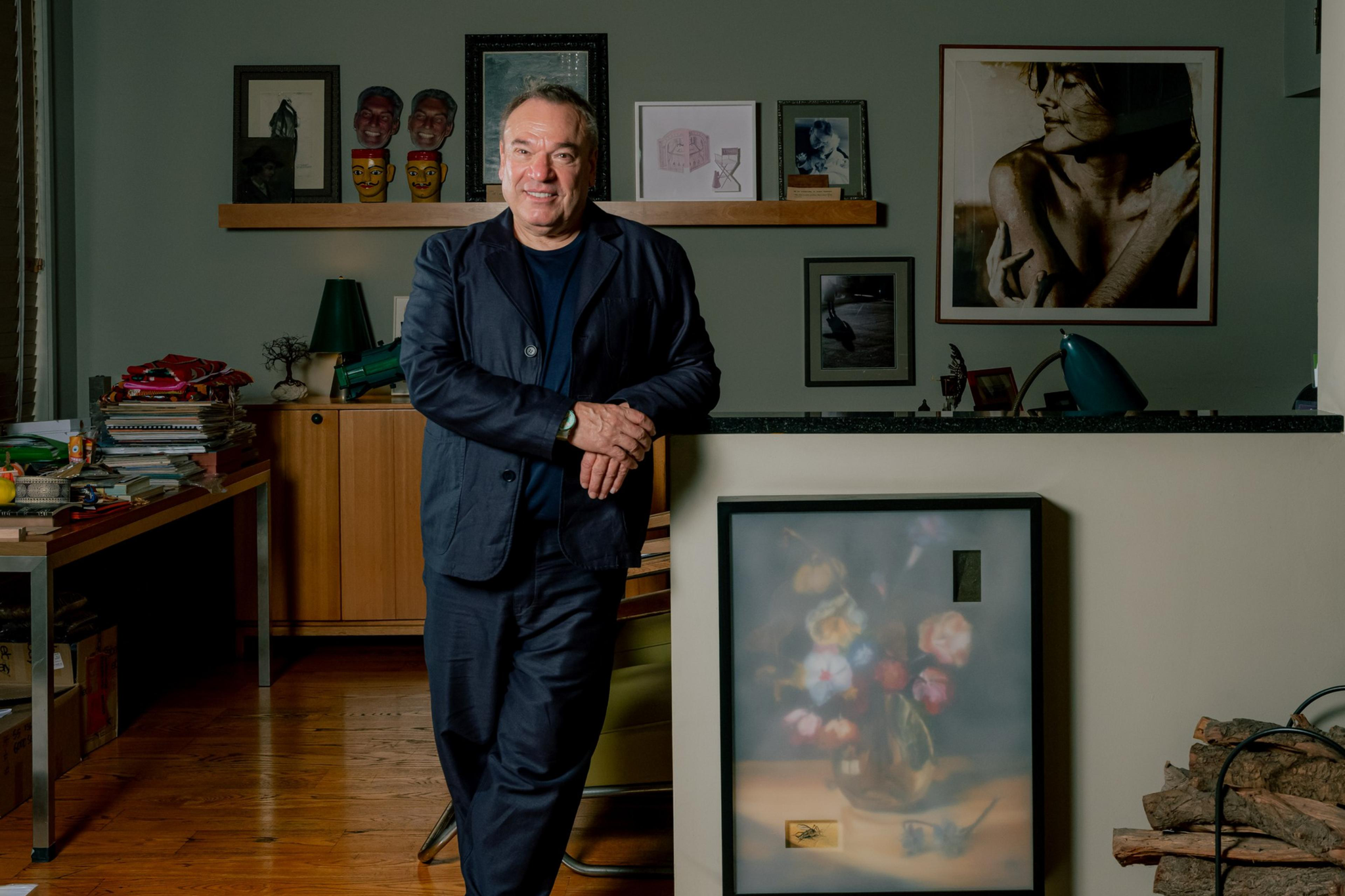 A man in a dark suit leans against a wall in a room with wooden floors, artworks on the wall, a table with stacks of items, and a pile of firewood.