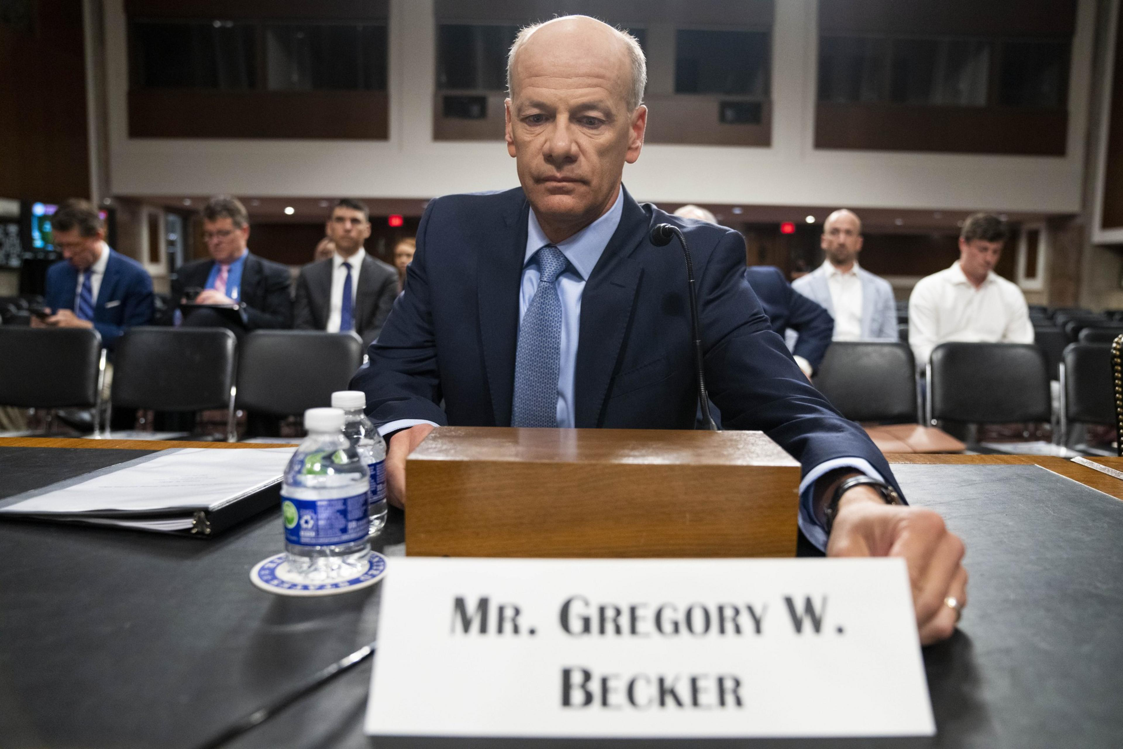 A man in a suit sits at a wooden desk with a microphone, nameplate, binder, and water bottles. Several people are seated behind him in a formal setting.