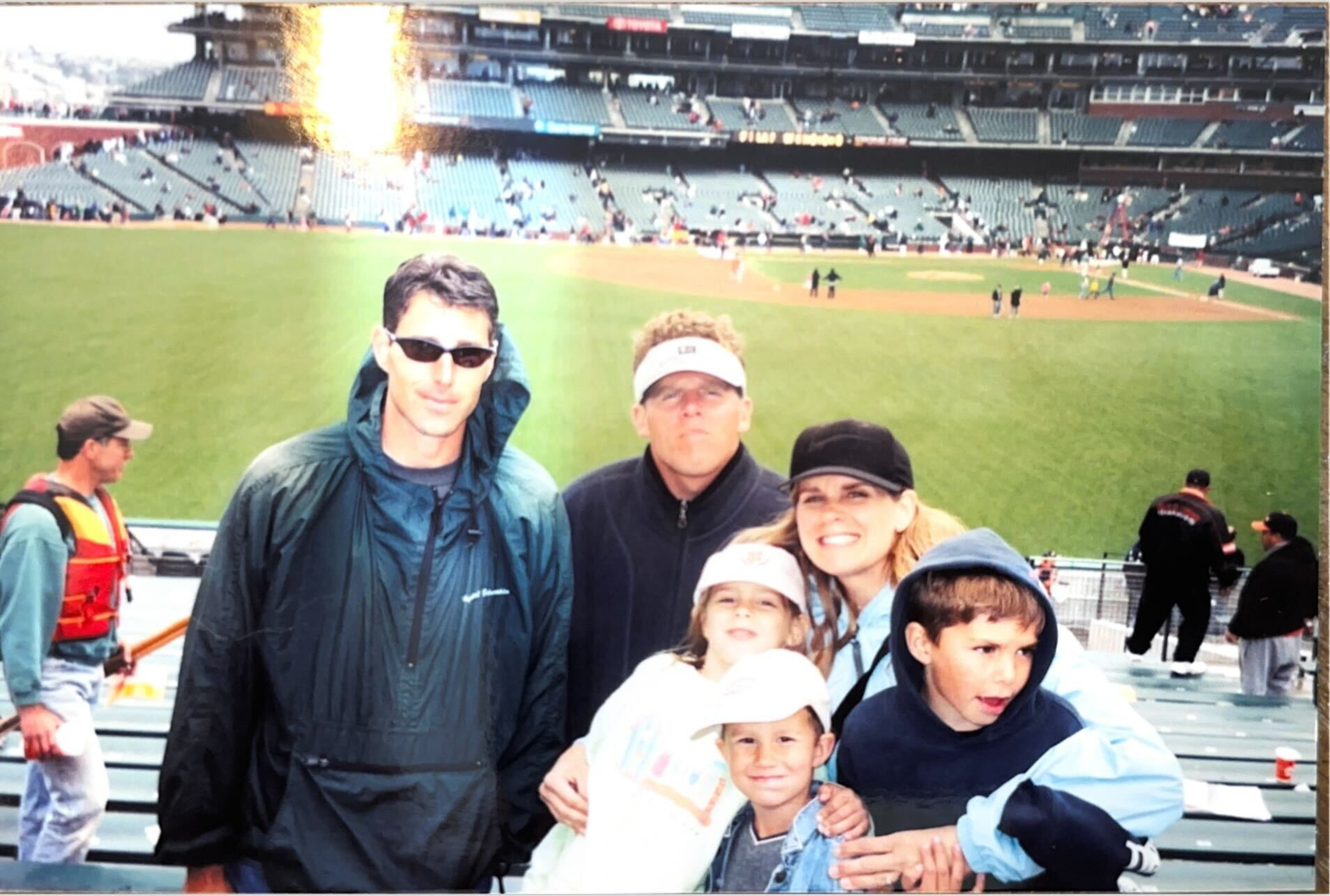 Two men, one woman, and three children pose smiling in front of a baseball field stadium with scattered spectators in the background.