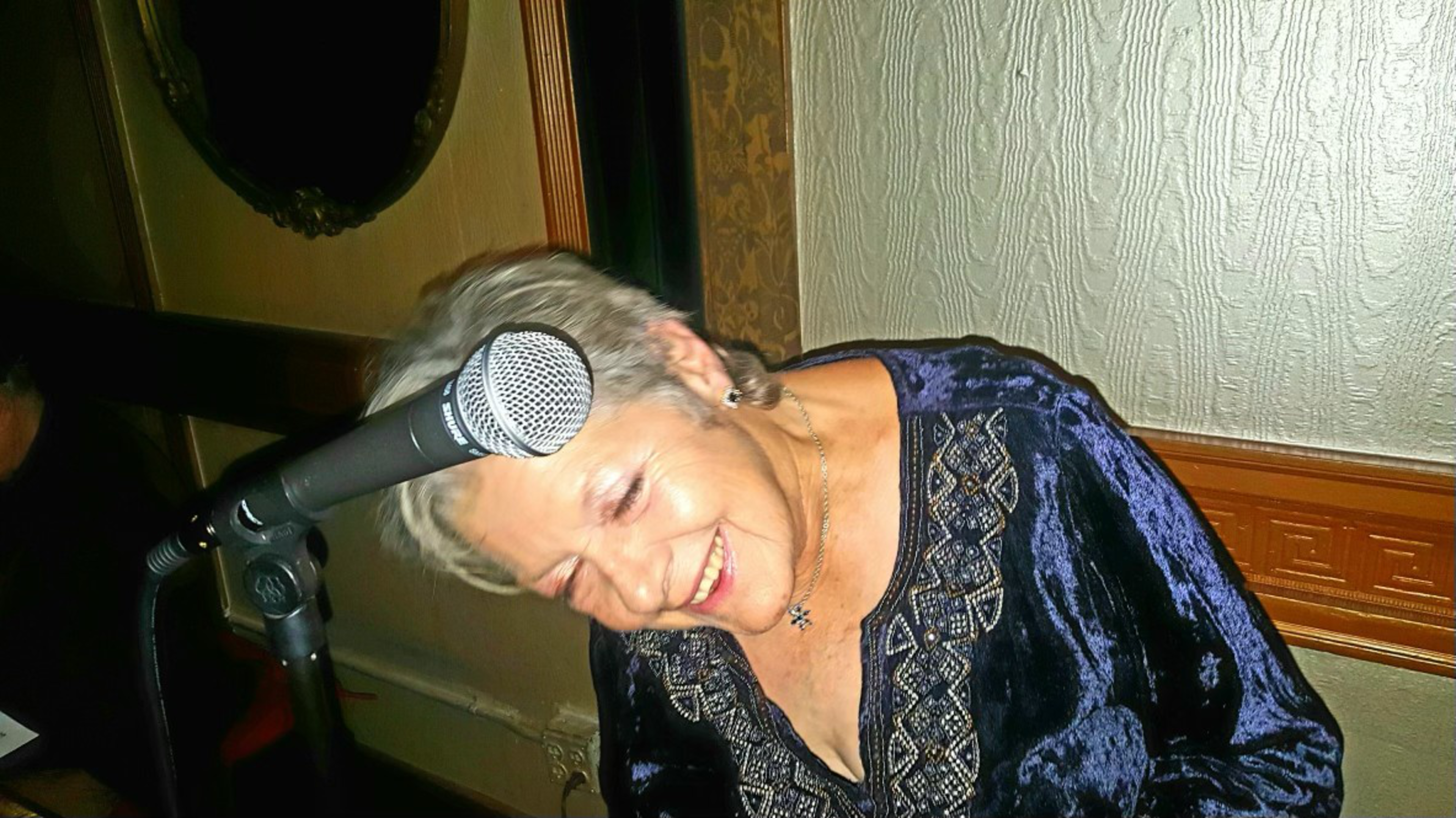 An elderly woman with short gray hair smiles warmly while leaning close to a microphone in a cozy, dimly lit room.