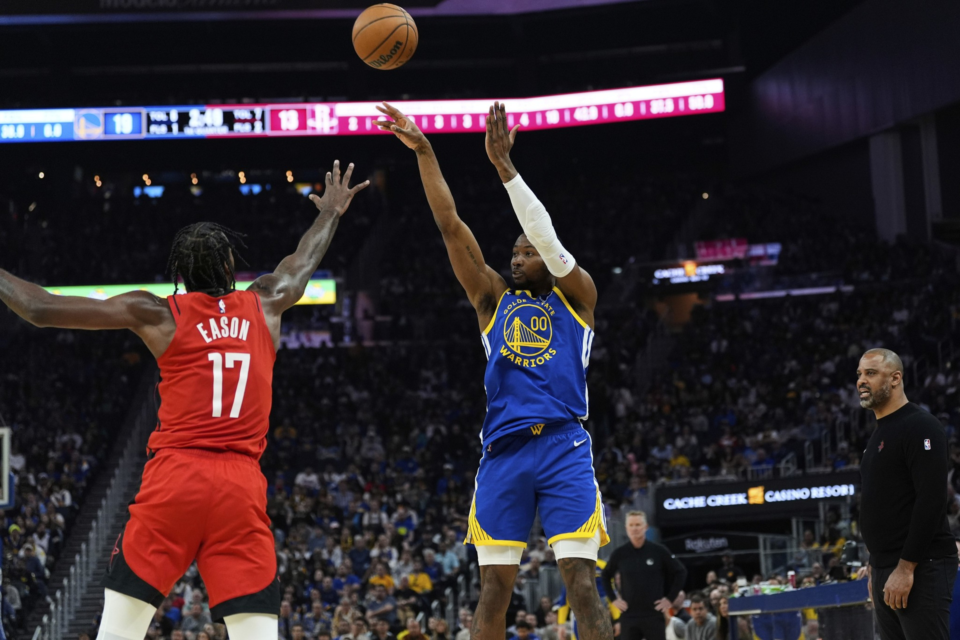 A basketball player in a blue jersey shoots the ball while defended by a player in a red jersey. A coach watches from the sideline in a crowded arena.