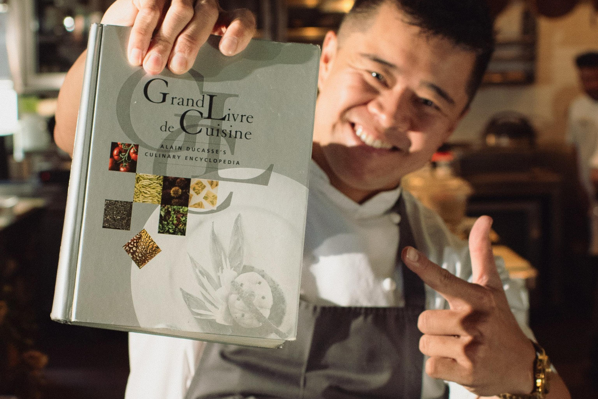 A person in a gray apron smiles and points while holding up a large cookbook titled "Grand Livre de Cuisine."