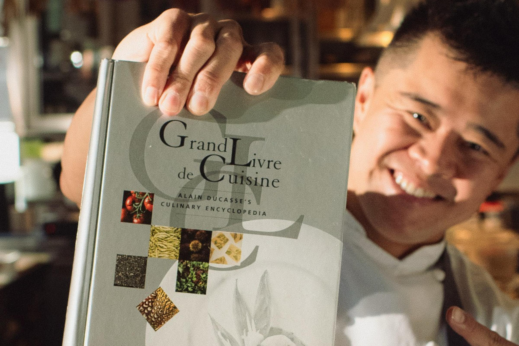 A person in a gray apron smiles and points while holding up a large cookbook titled "Grand Livre de Cuisine."