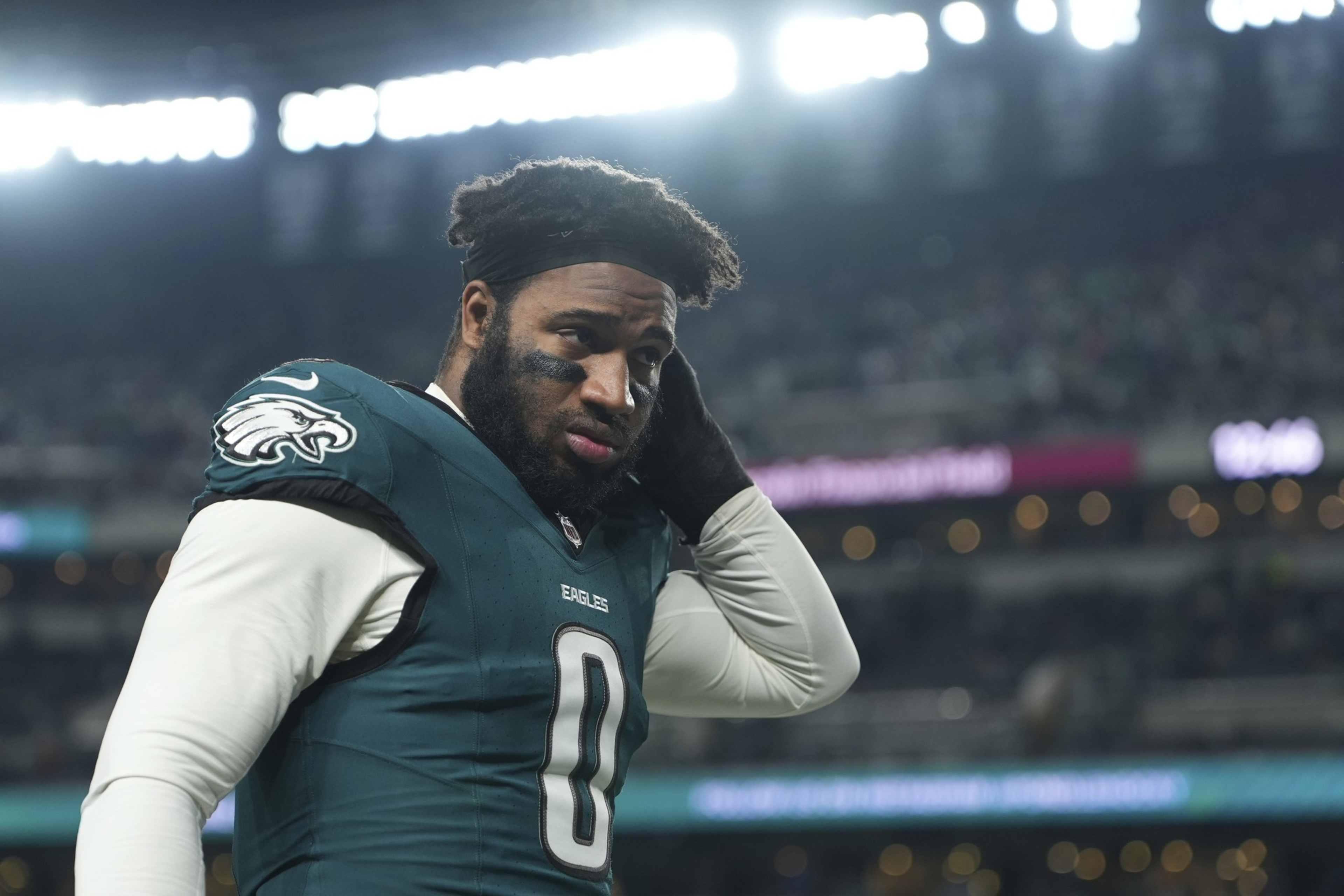 A football player in a green and white uniform with the number 0 adjusts his hair under stadium lights. He has black eye paint and looks focused.