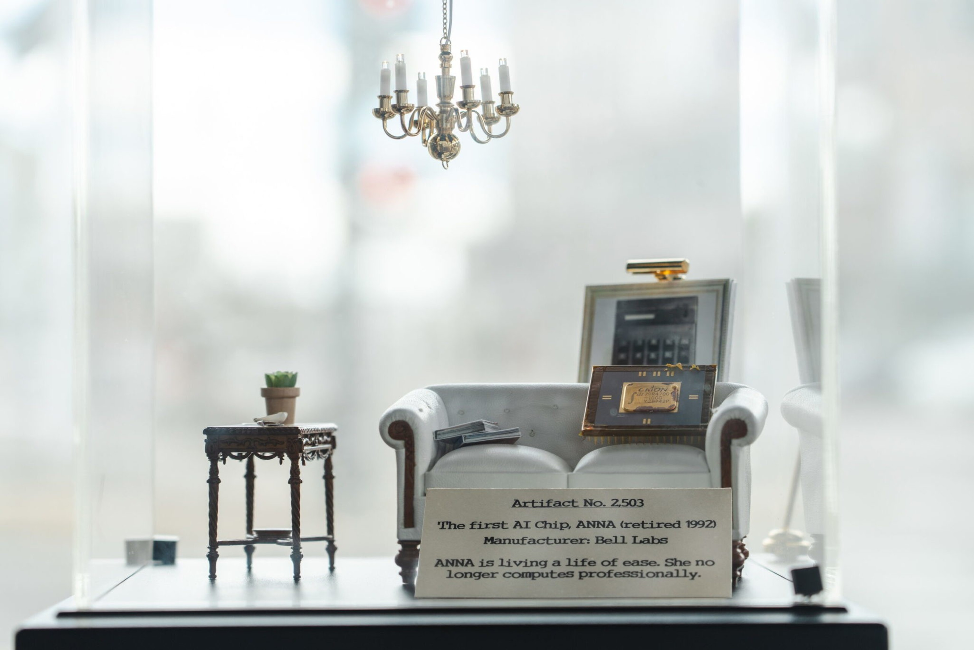 A miniature sofa with a framed AI chip rests beside a small table with a potted plant, under a hanging chandelier, displayed with a descriptive plaque.