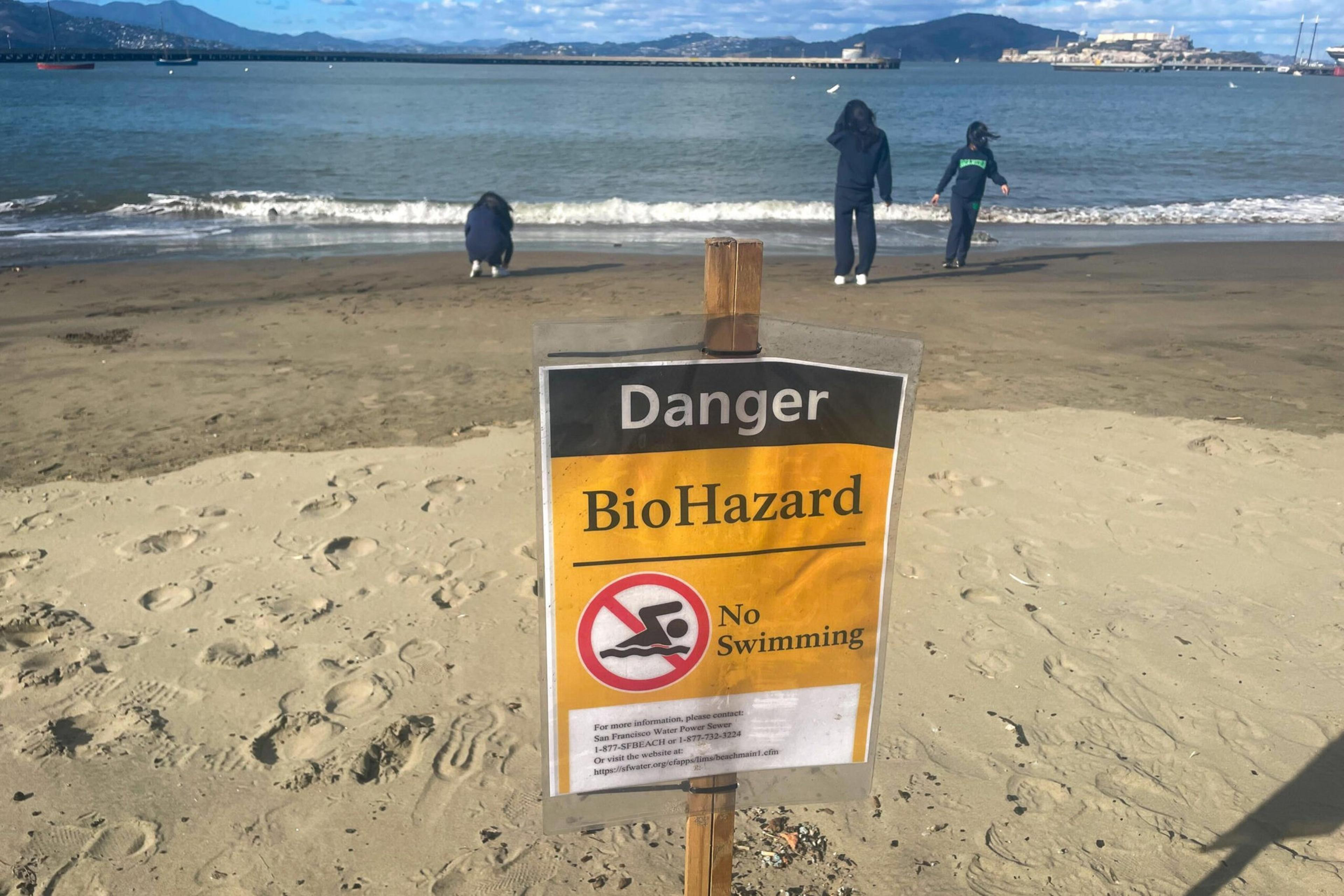 A beach with a biohazard warning sign stating "Danger BioHazard No Swimming" is posted, while three people stand near the water's edge with mountains in the background.
