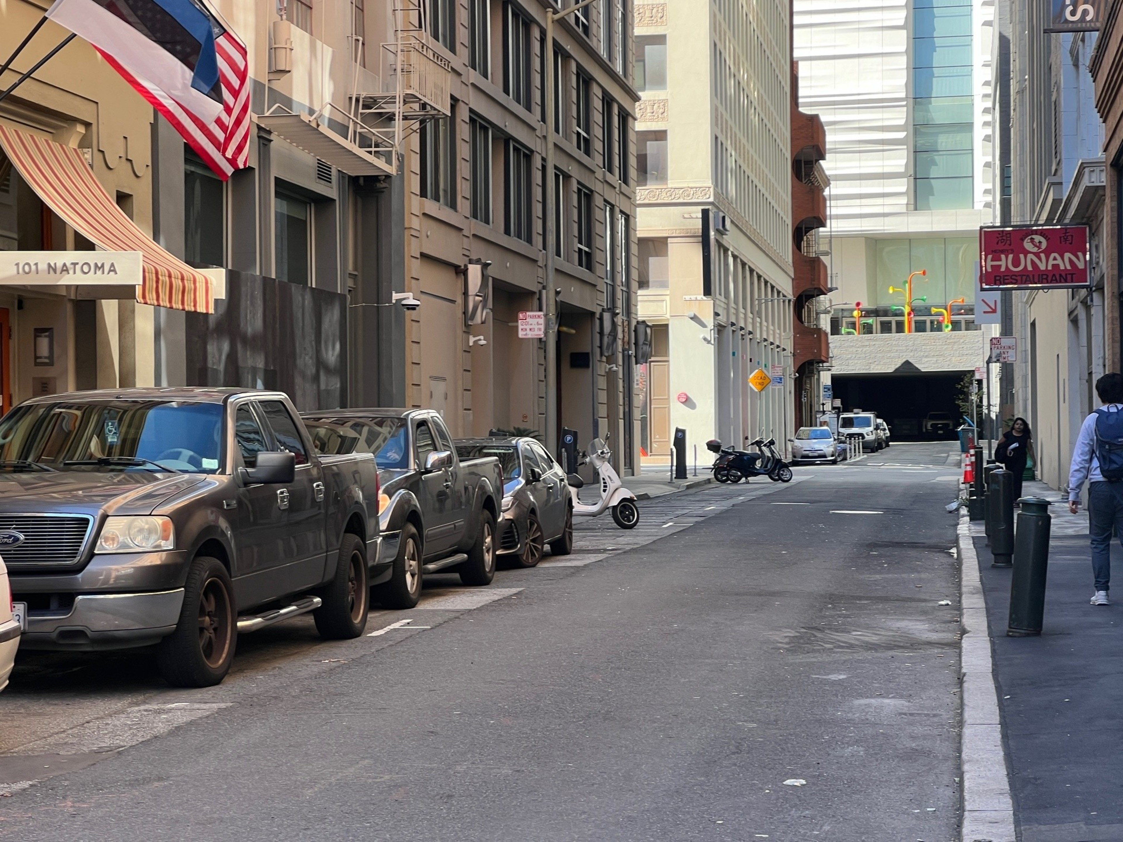 Urban street with parked cars, scooters, pedestrians, flag, and a restaurant sign.