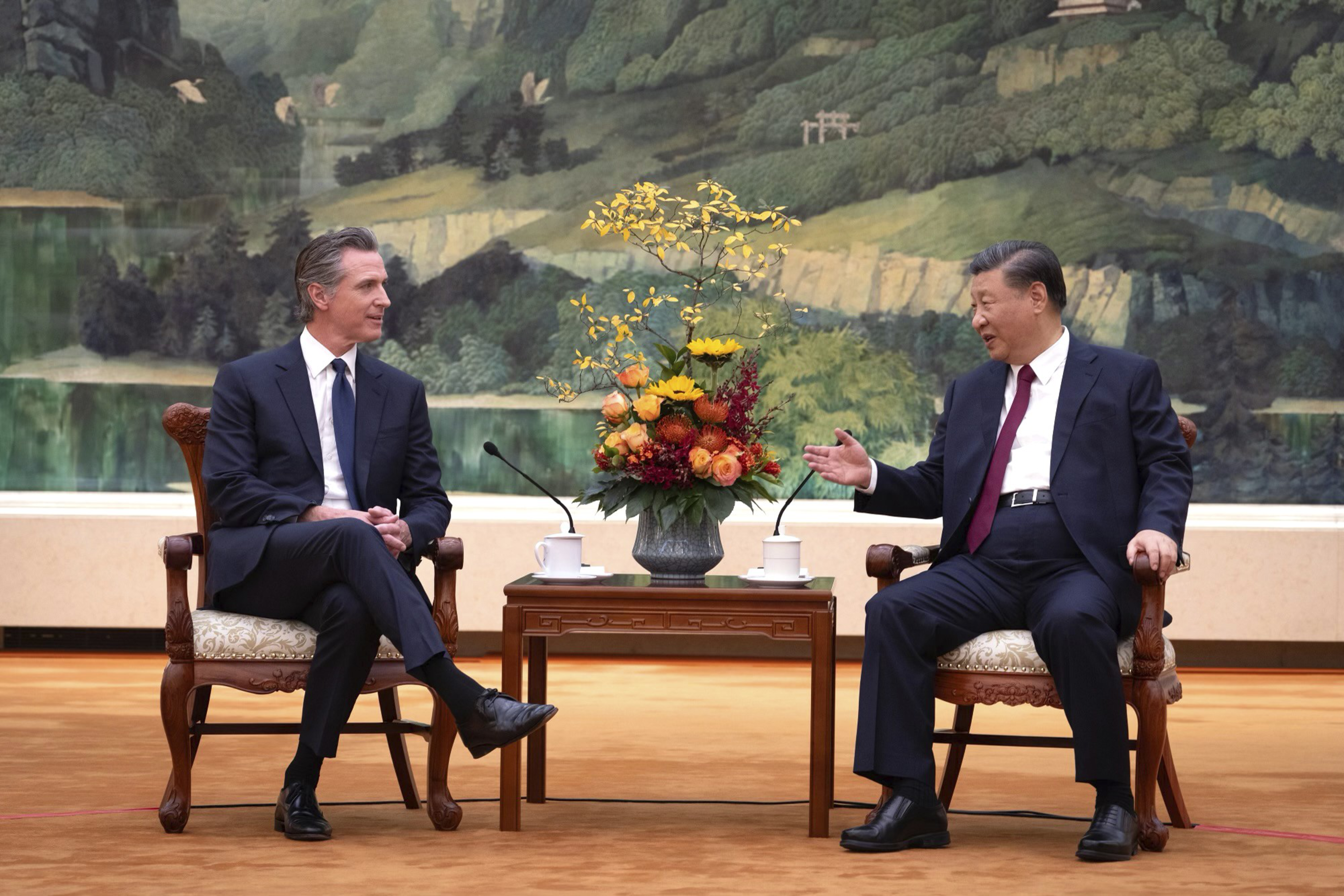 Two men in suits sit in ornately carved chairs with a small wooden table and a flower arrangement between them, engaged in conversation against a painted backdrop.