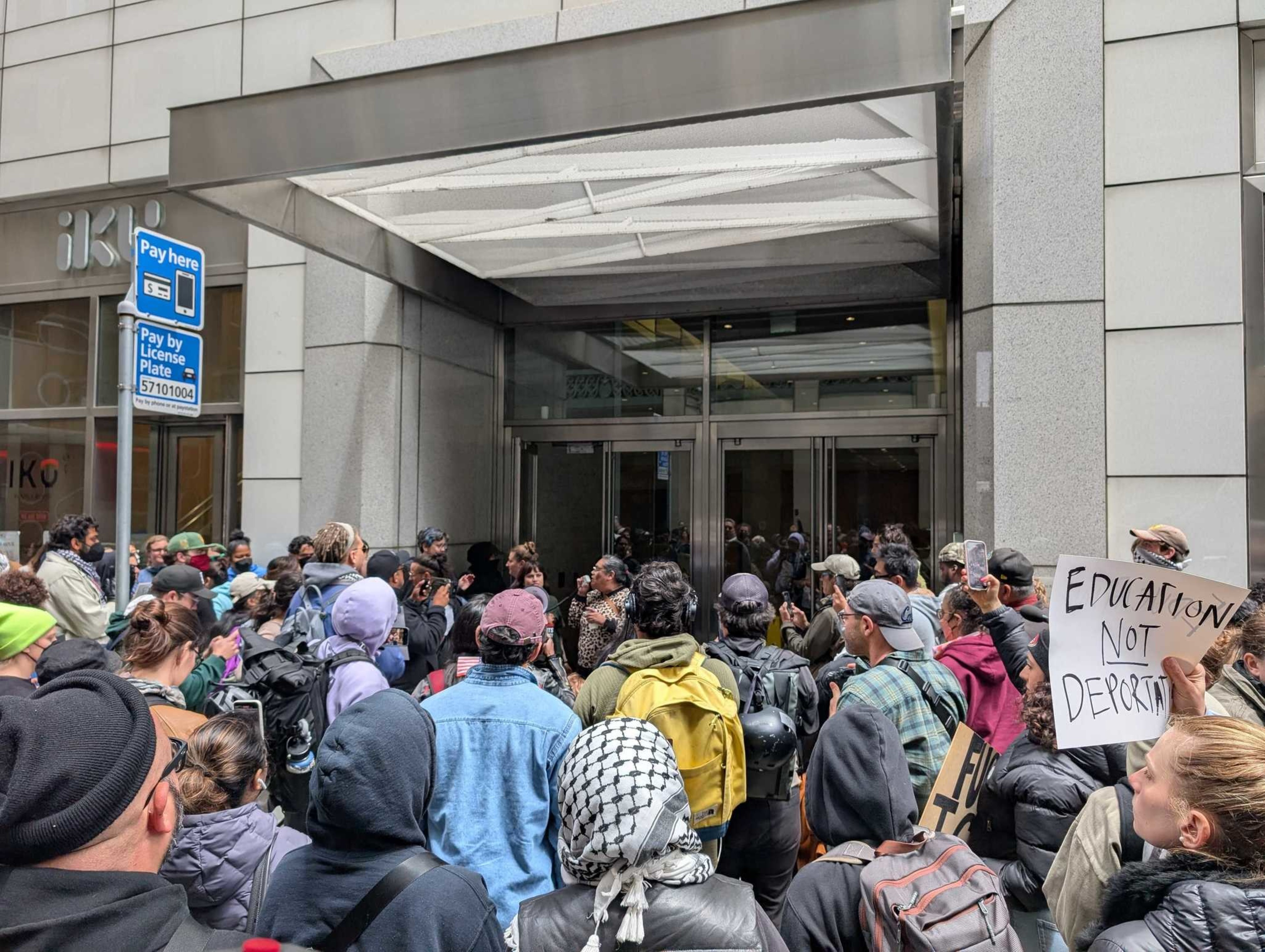 A large group of people gathers outside a building entrance, holding signs with messages like &quot;Education Not Deportation,&quot; suggesting a protest or demonstration.
