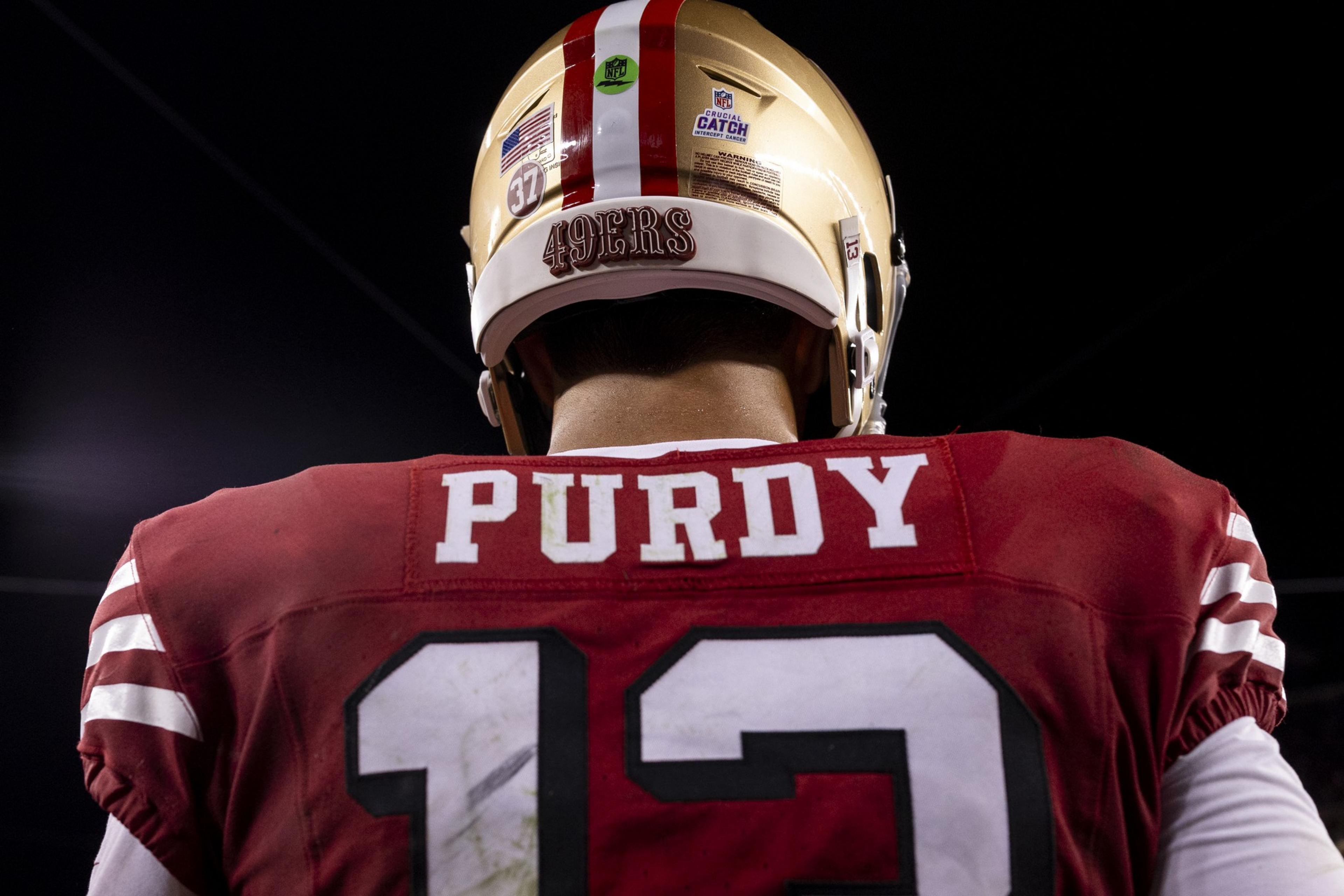 A football player in a red jersey with the name &quot;Purdy&quot; and number 13, wearing a gold helmet with &quot;49ERS&quot; and an NFL logo, faces away in a dimly lit setting.