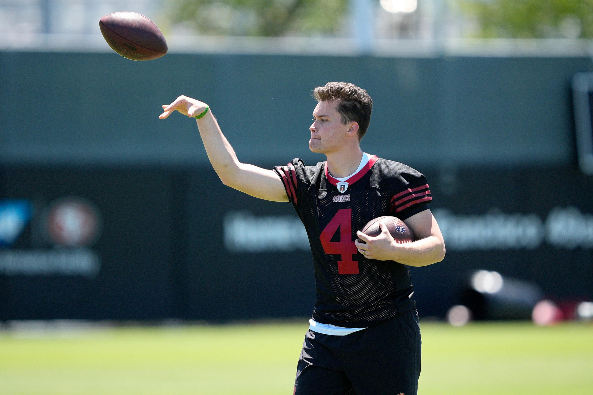 A football player wearing a black jersey with number 4 is throwing a football while holding another football in his left arm on a sunny field.