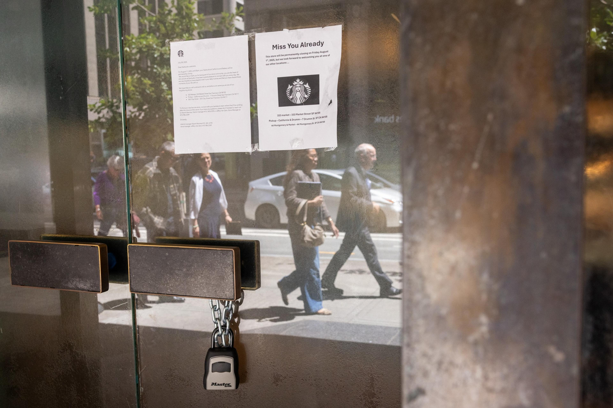 A locked glass door with a padlock has two signs taped on it, reflecting pedestrians and cars passing outside on a sunny day.