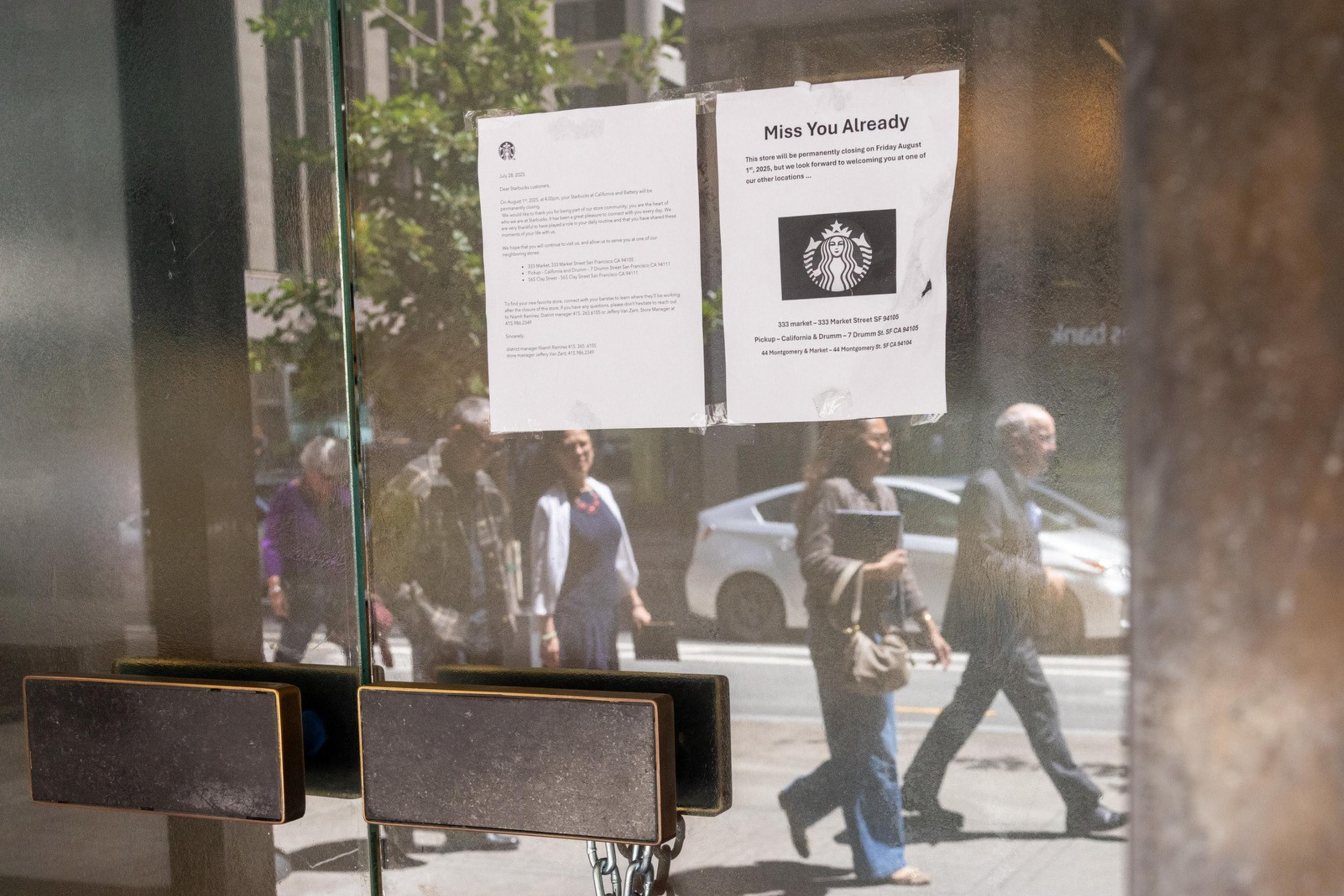 A locked glass door with a padlock has two signs taped on it, reflecting pedestrians and cars passing outside on a sunny day.