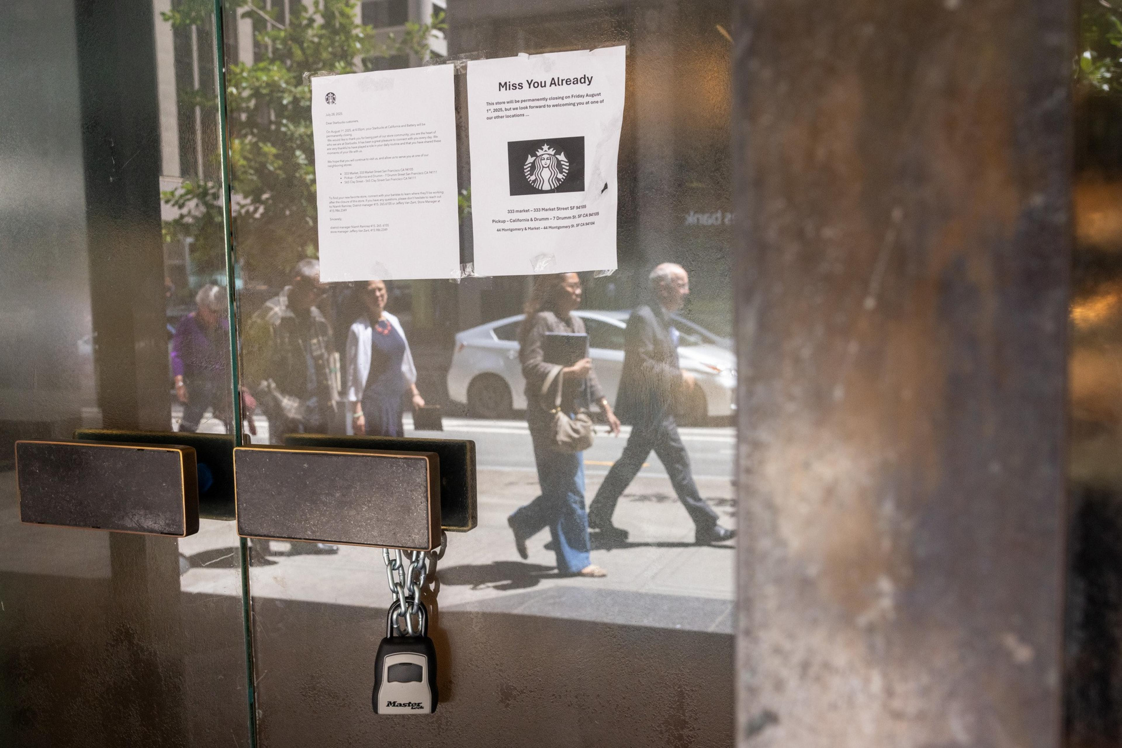 A locked glass door with a padlock has two signs taped on it, reflecting pedestrians and cars passing outside on a sunny day.